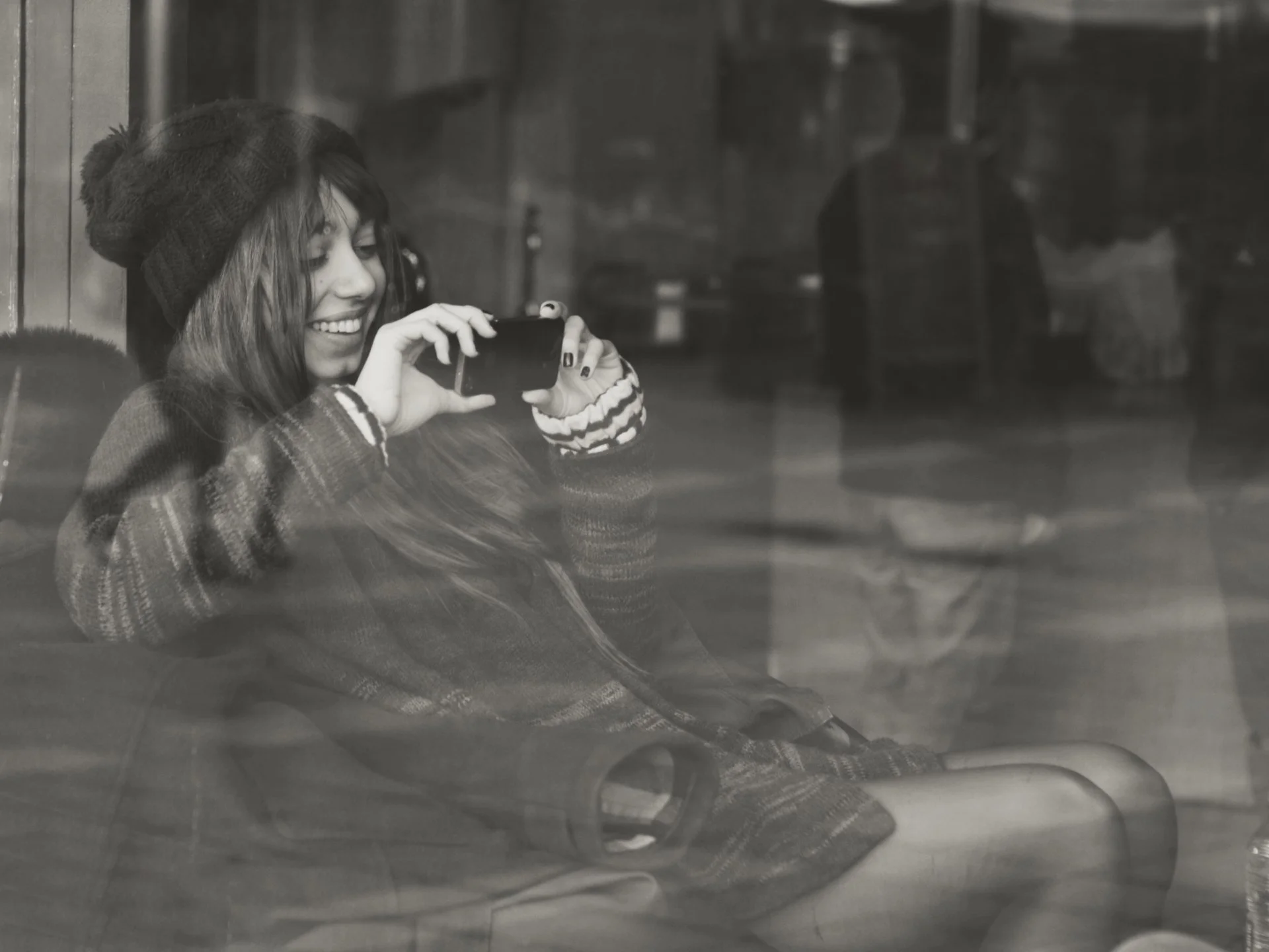 A young woman with long hair, wearing a beanie and striped sweater, is sitting and smiling while looking at her phone. She is seen through a reflective surface, possibly a window.