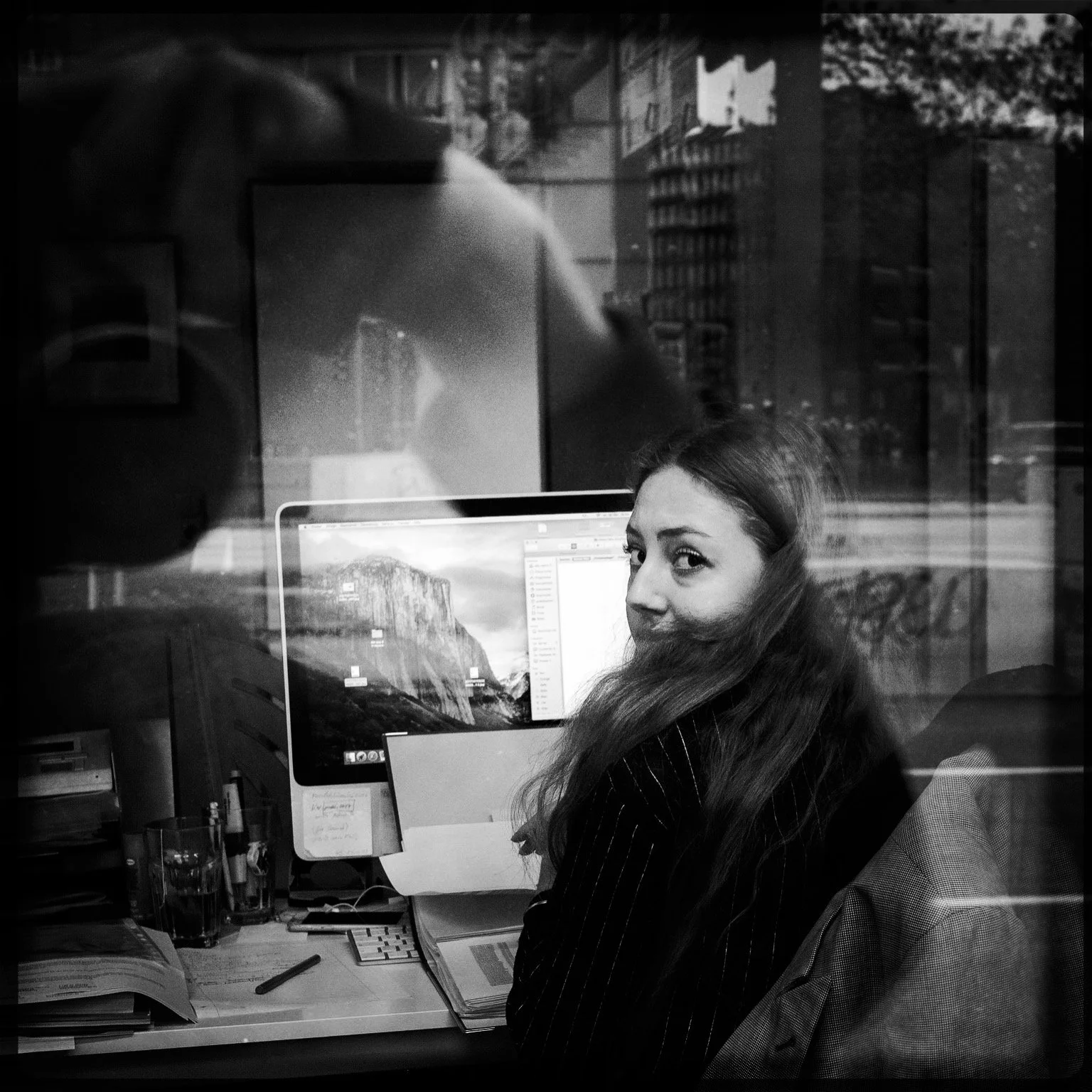 A woman with long hair sitting at a desk with computer monitors, papers, and pens, looking through a glass window.