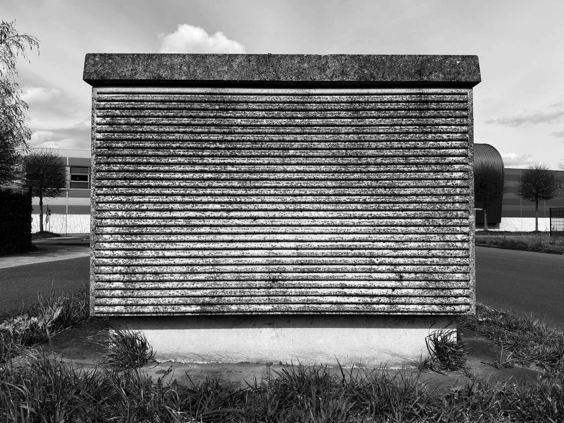 Black and white exterior view of a modern building with a textured ribbed wall and a curved section on the right, surrounded by grass and trees.