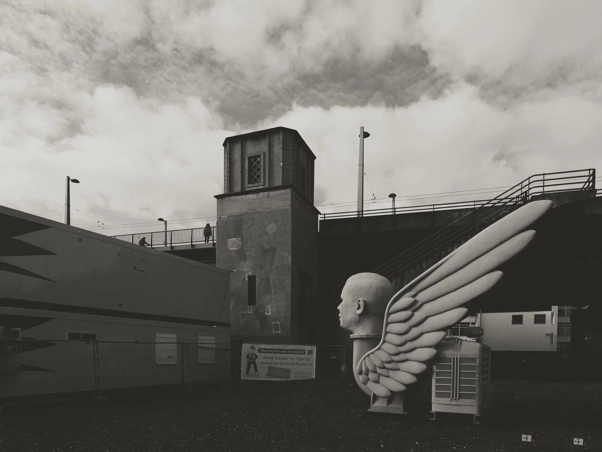 Black and white photo of an urban scene with a building, a bridge, and decorative wing sculptures. There are a few pedestrians on the bridge and utility poles beneath a cloudy sky.