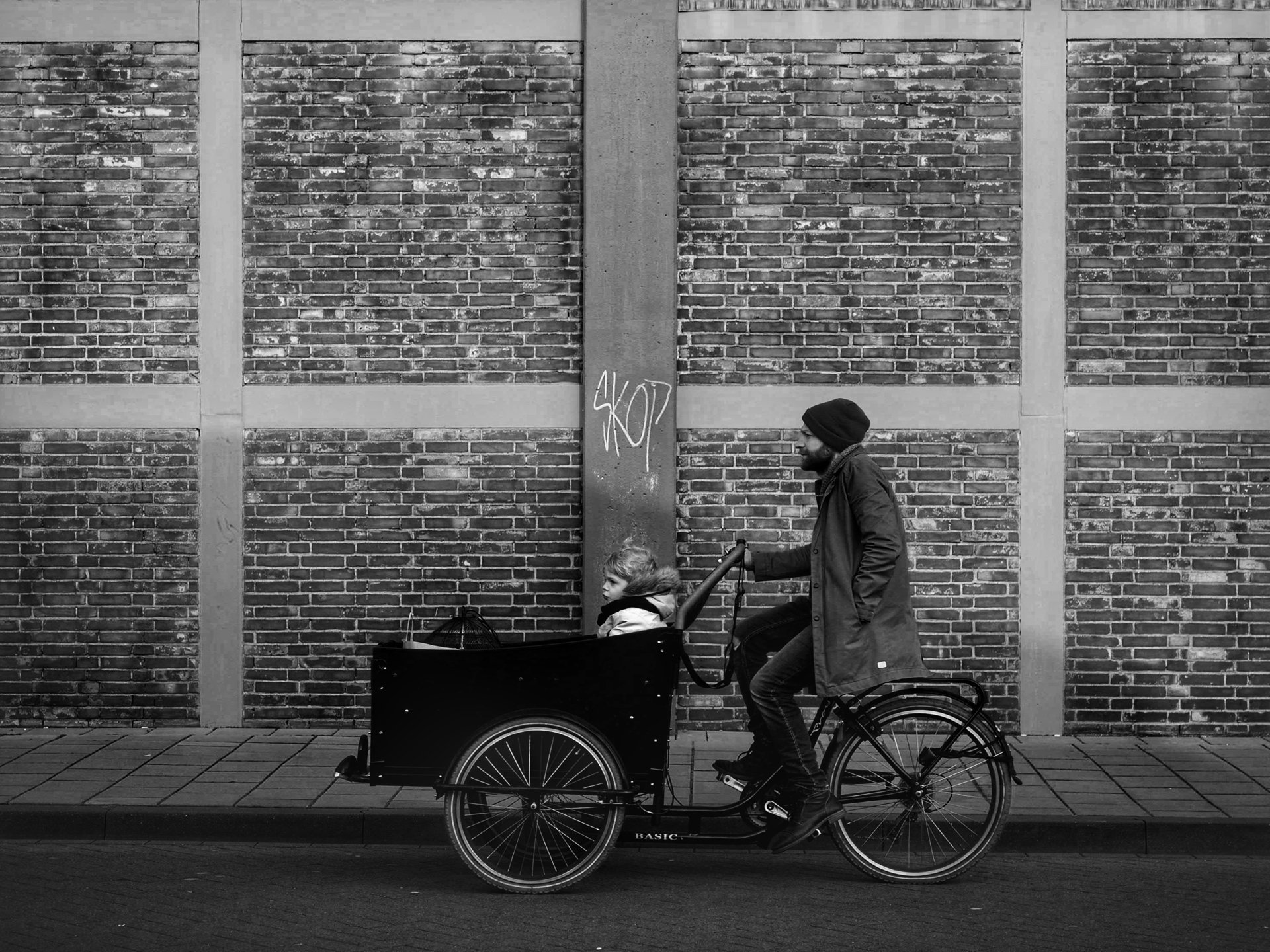 A man in a jacket and beanie riding a cargo bicycle with a child in the front cargo area, against a brick wall with graffiti reading 'SKOP' in black spray paint.