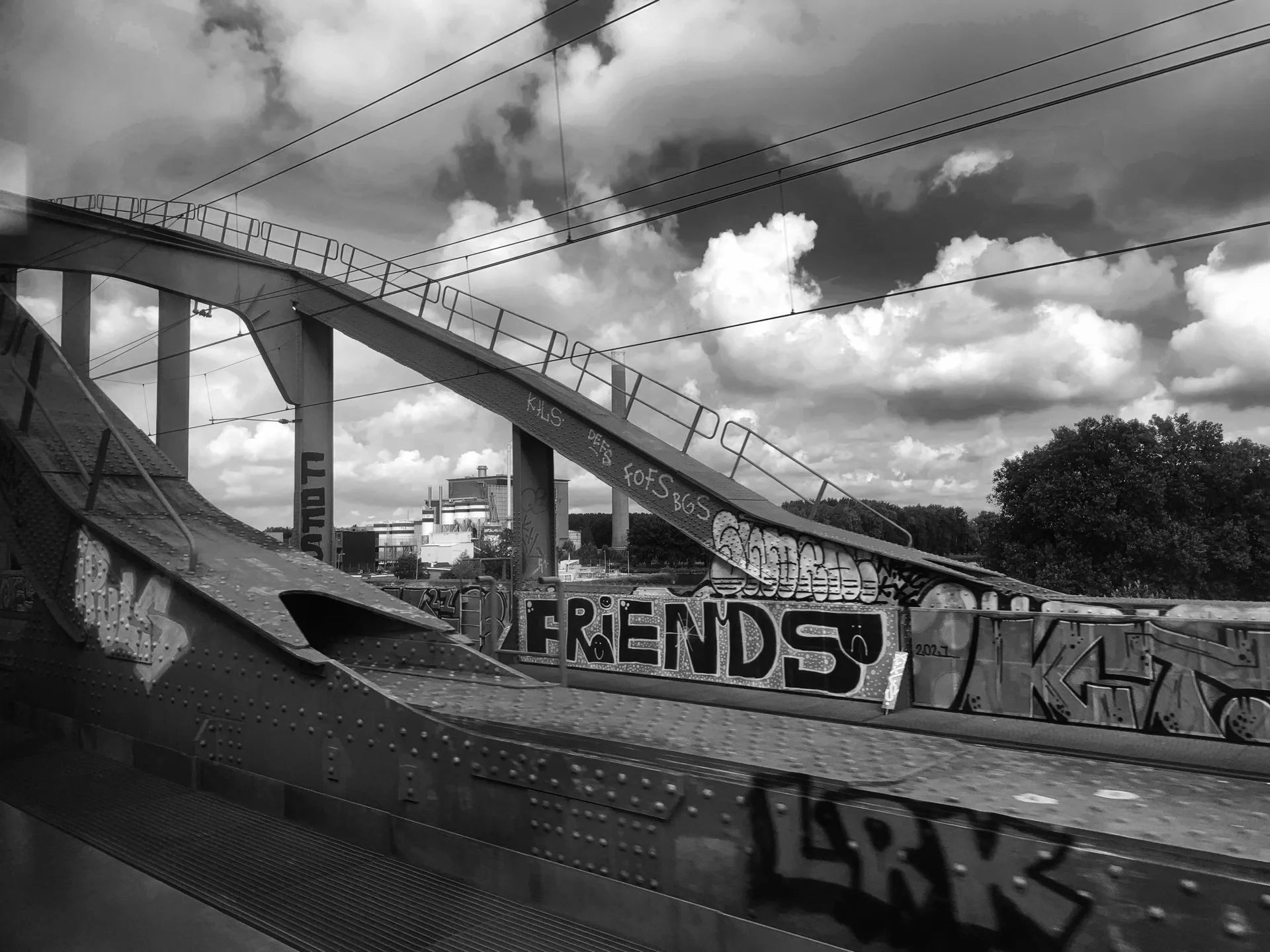 Black and white photo of a graffiti-covered bridge or ramp with clouds and a cityscape in the background.