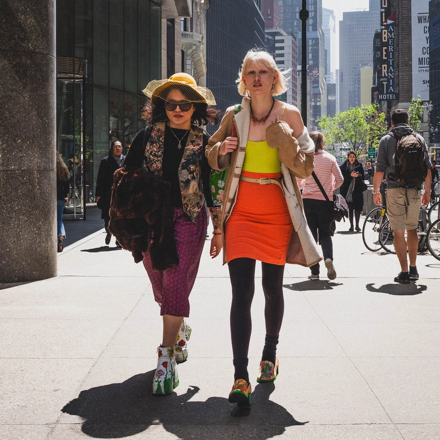 Two women walking on a city sidewalk during daytime, surrounded by pedestrians and tall buildings. One woman wears a large hat and sunglasses, the other has short, blonde hair and a colorful outfit.