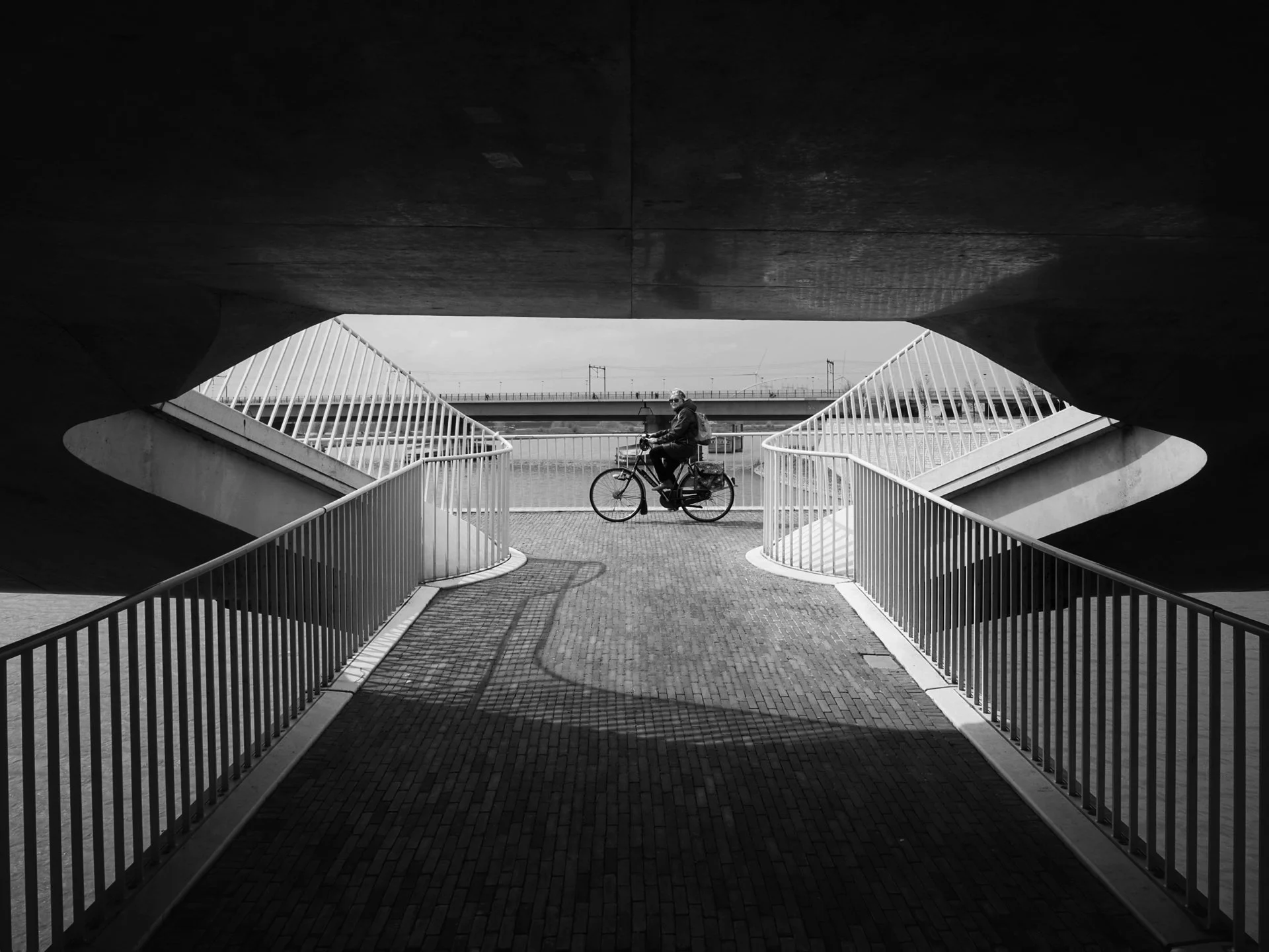 A person riding a bicycle under a modern bridge with sleek, curved railings and a paved walkway.