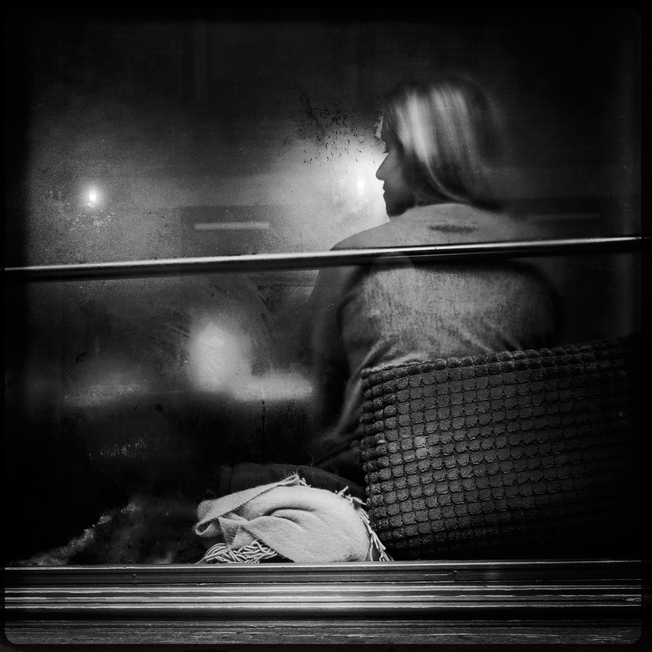 A woman sitting alone on a train or bus at night, gazing out the window, with condensation on the glass and faint lights outside.