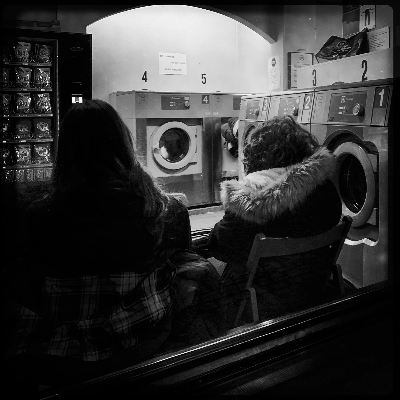 Two women sit in front of washing machines and dryers in a laundromat, viewed through a window.