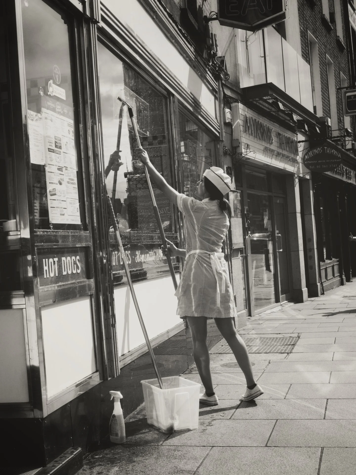 A woman in a striped apron and a chef's hat uses a squeegee to clean the glass window of a storefront featuring a sign for hot dogs. She is on a city sidewalk with various storefronts visible in the background.