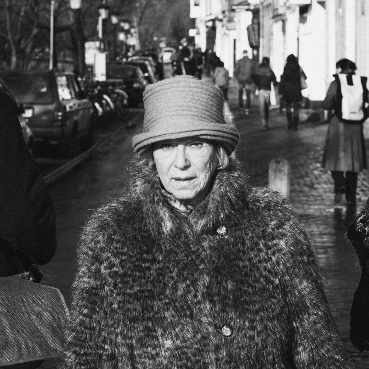 A black-and-white photo of an elderly woman with light skin and a serious expression, wearing a large fur coat and a wide-brimmed hat, walking on a cobblestone street with parked cars and pedestrians in the background.
