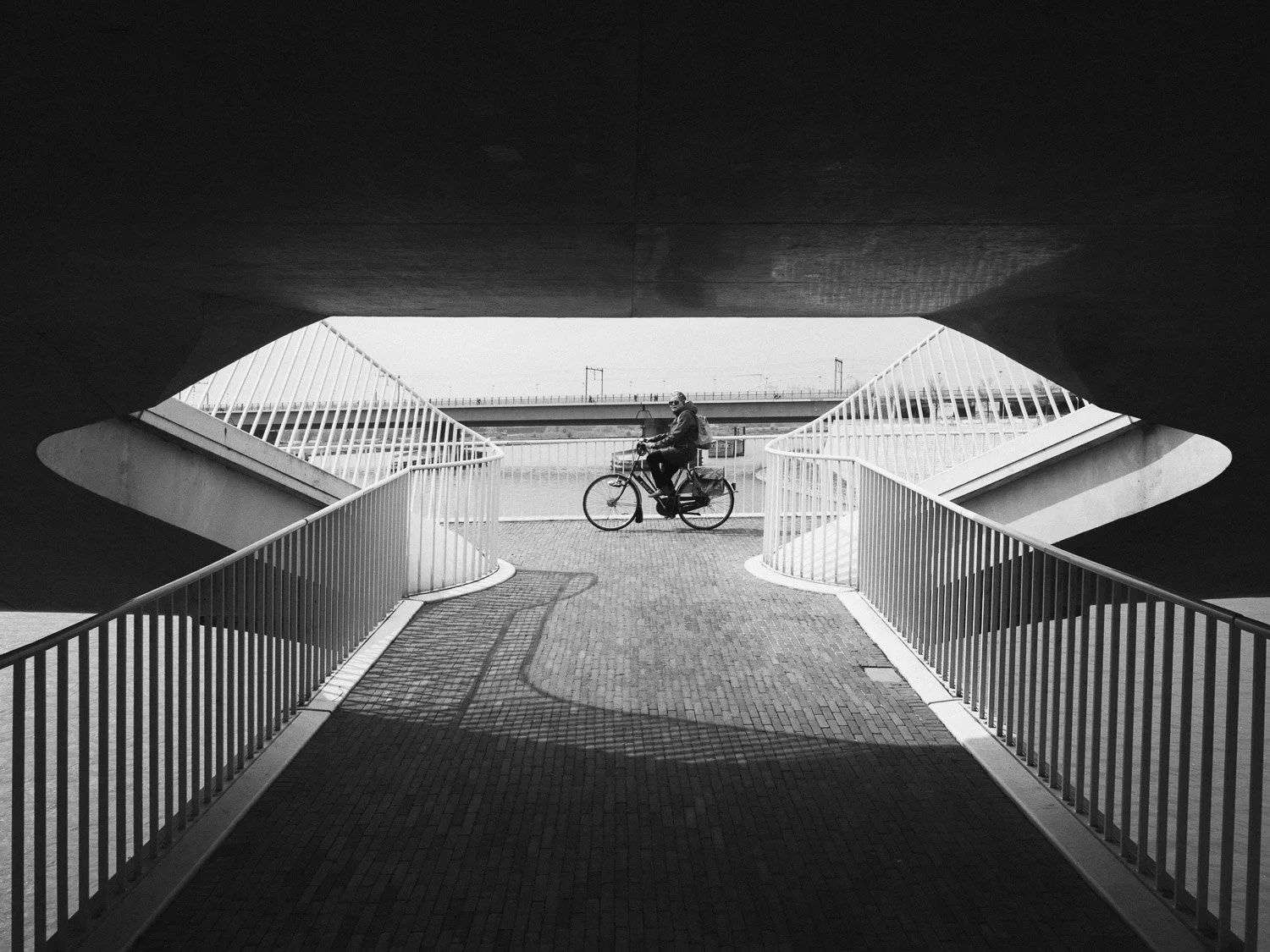 Black and white photo of a person riding a bicycle on a bridge, viewed from beneath an overpass with arching railings on either side.