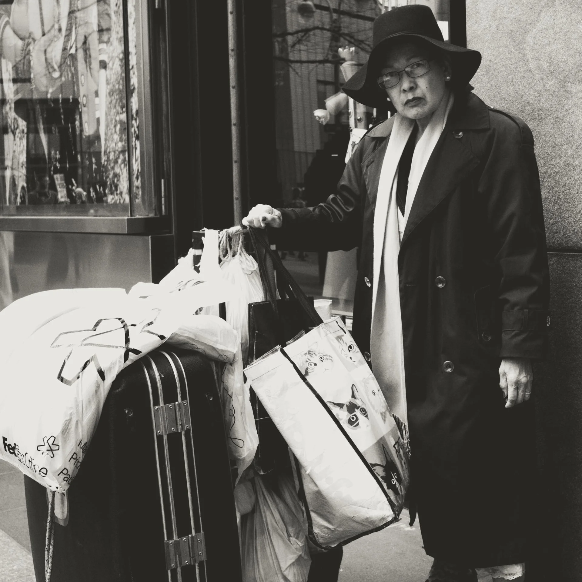 A woman wearing glasses, a wide brim hat, and a trench coat is standing next to a vending machine, holding shopping bags in one hand, with a serious expression.
