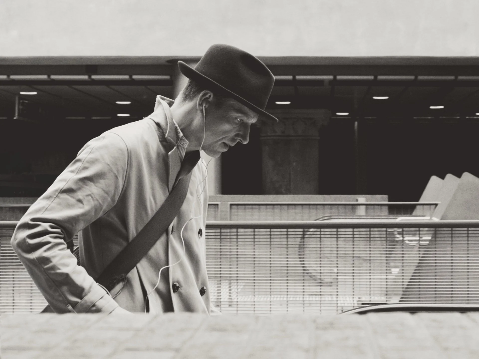 Black and white photo of a man in a coat and hat, looking down and listening to earphones, while carrying a bag, in an urban setting.