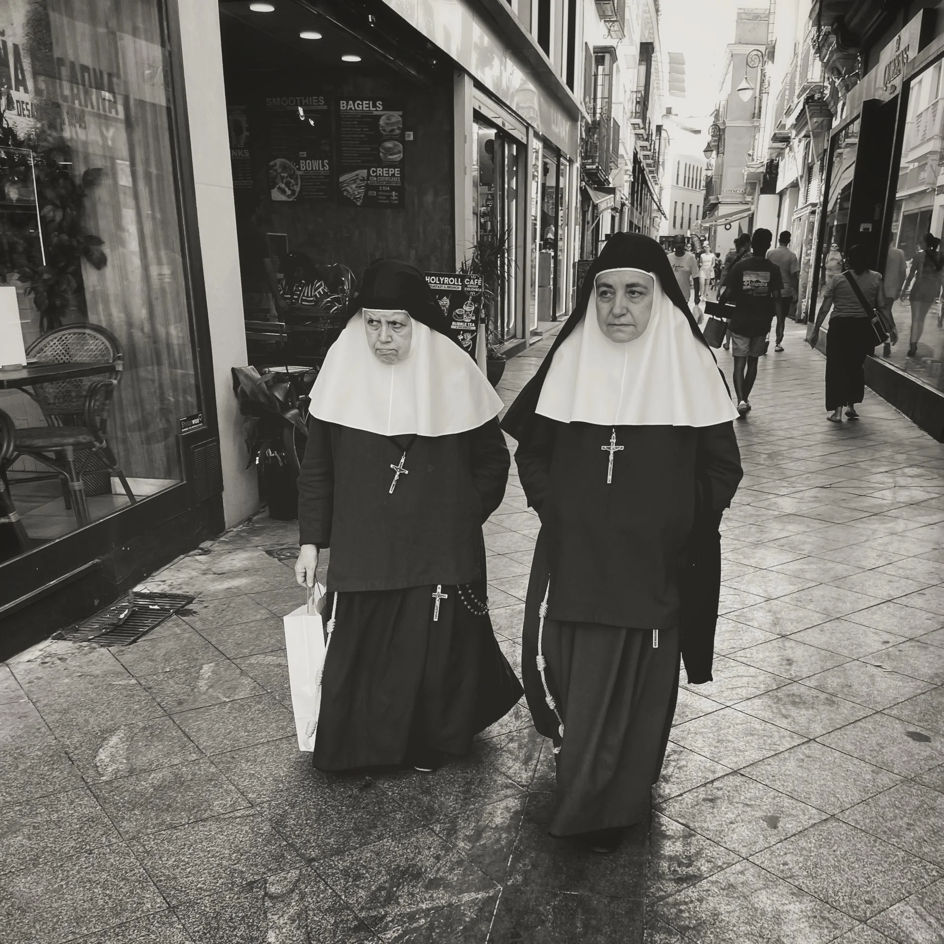 Two nuns wearing traditional black and white habits walk down a city sidewalk lined with shops and pedestrians.