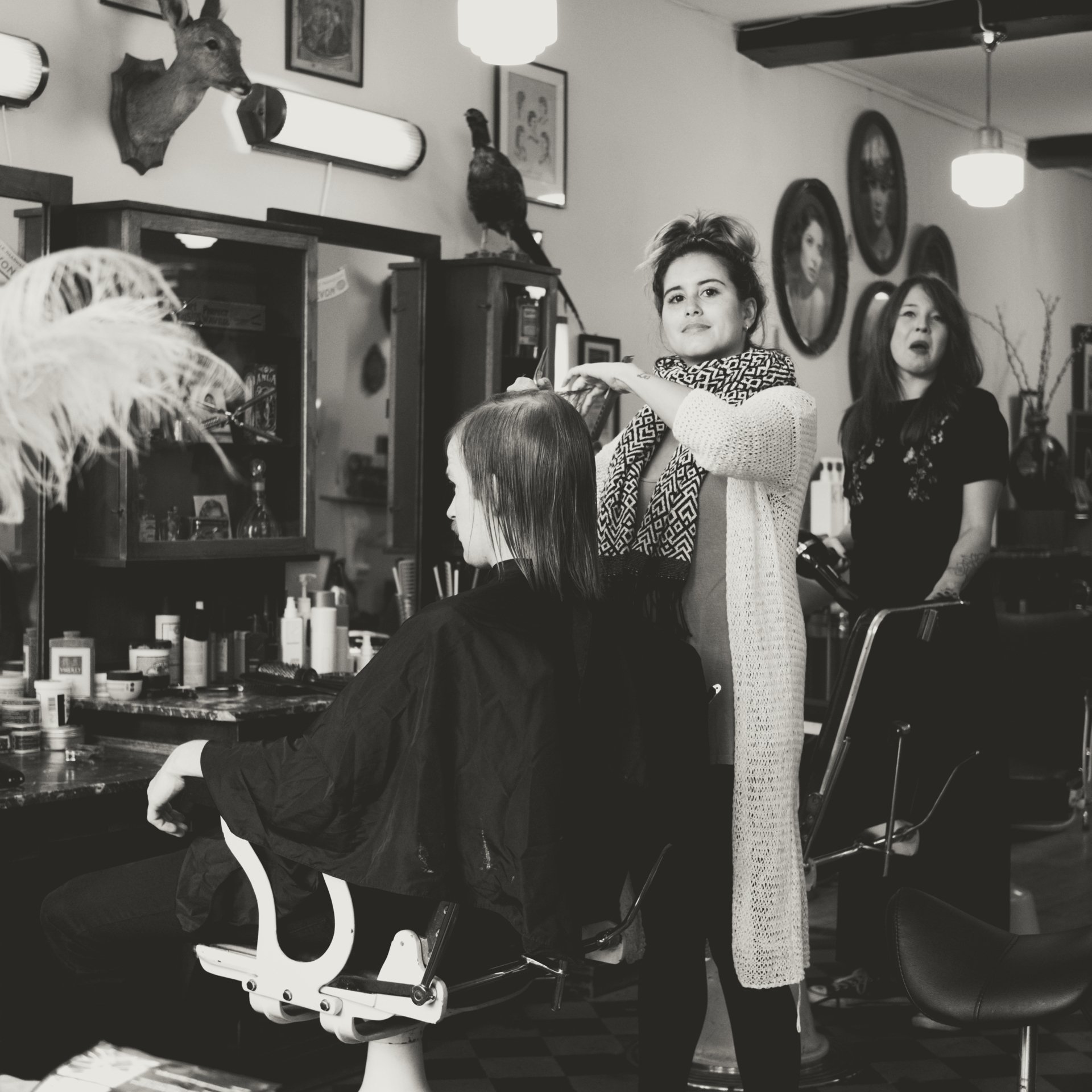 A woman getting her hair done at a salon, with a deer head mounted on the wall and a portrait of a woman in the background.