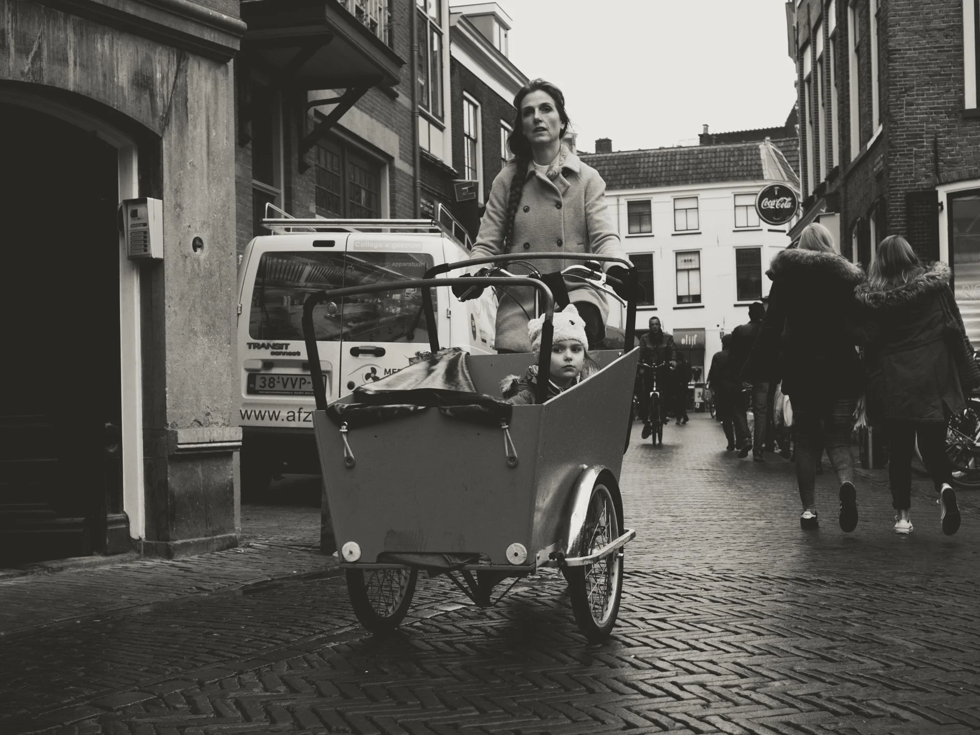 Black and white street scene with a woman riding a cargo bicycle with a child in the front basket. Pedestrians walk on the sidewalk, and there are buildings and a white van in the background.