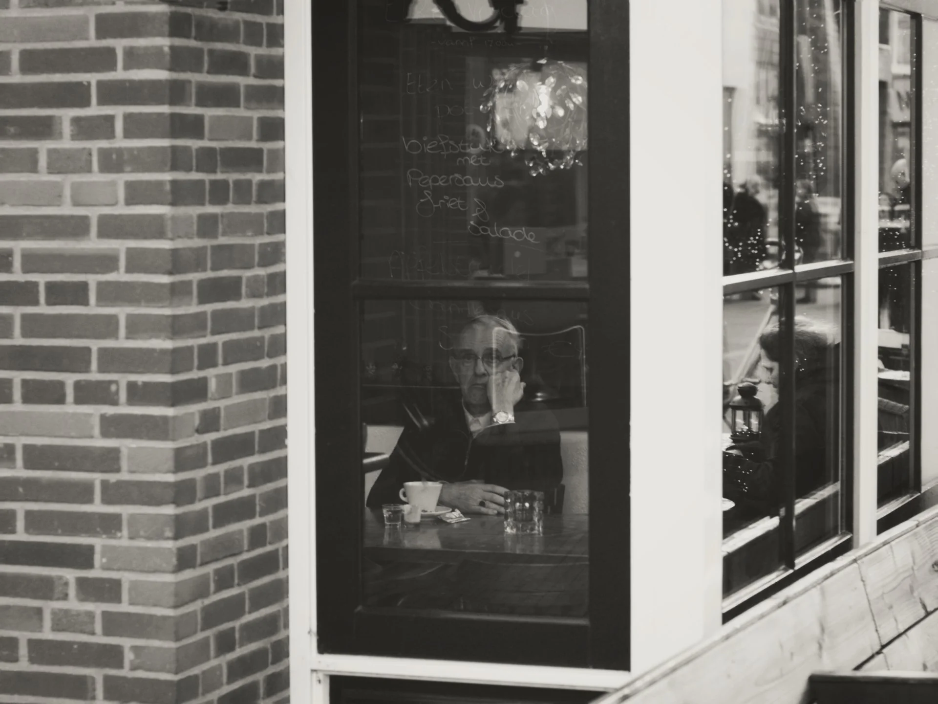 A man in glasses sitting alone at a table inside a cafe or restaurant, looking at his phone, seen through a glass window from outside. The table has a cup, a glass of water, and some papers. Behind the man, there are faint reflections of Christmas li