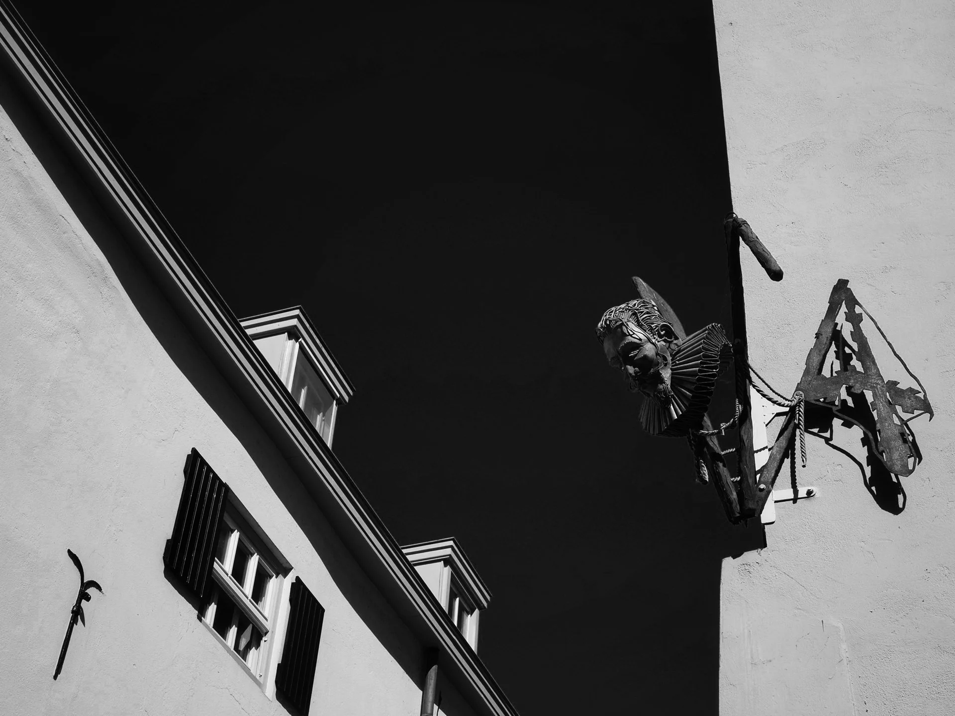 A black and white photo of the exterior of a building showing three small windows with black shutters, a roof edge, and a decorative sculpture resembling a clown head with a ruffled collar, mounted on a wall and attached to a metal frame.