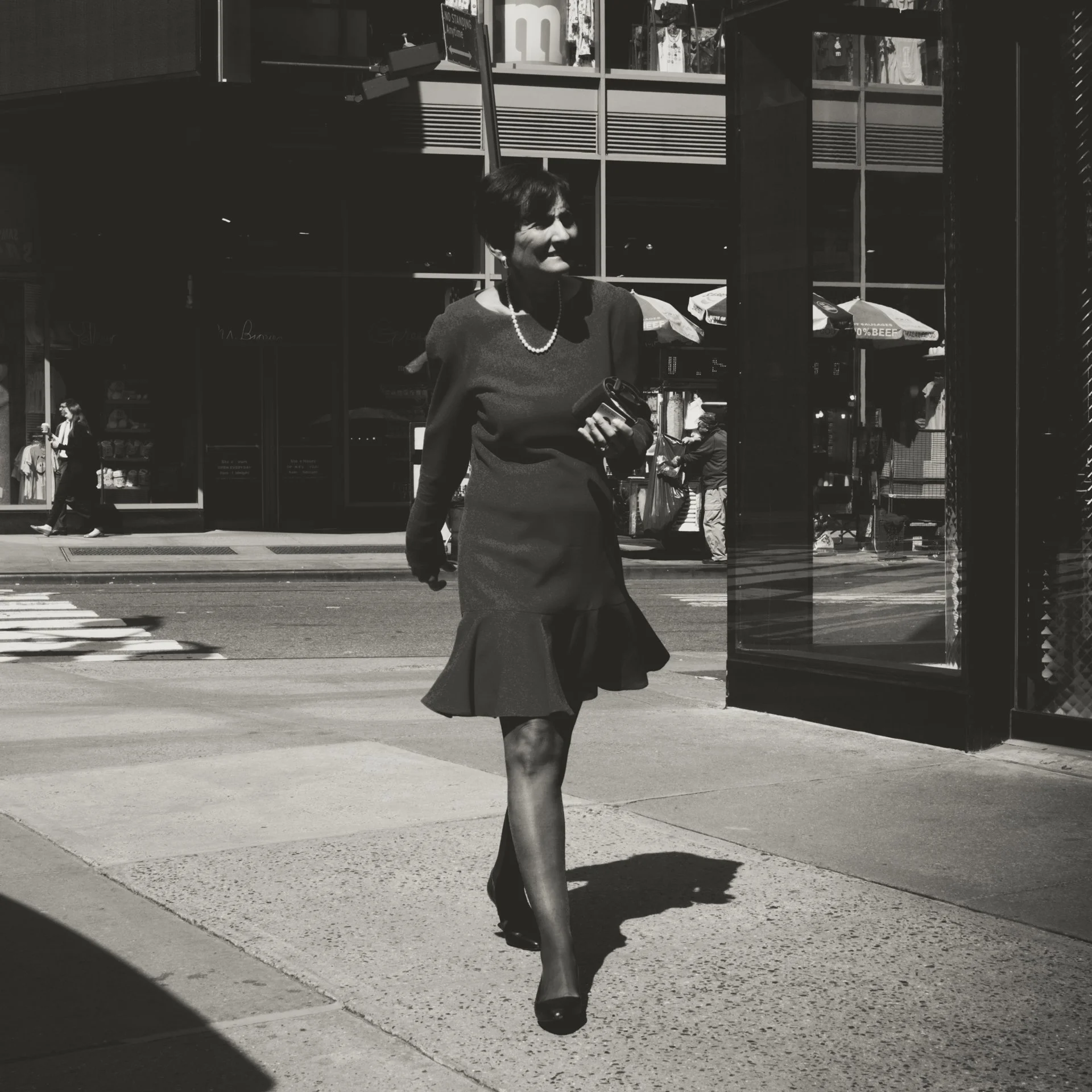 A woman crossing the street in a business outfit, walking past a phone booth, with a city retail store in the background.
