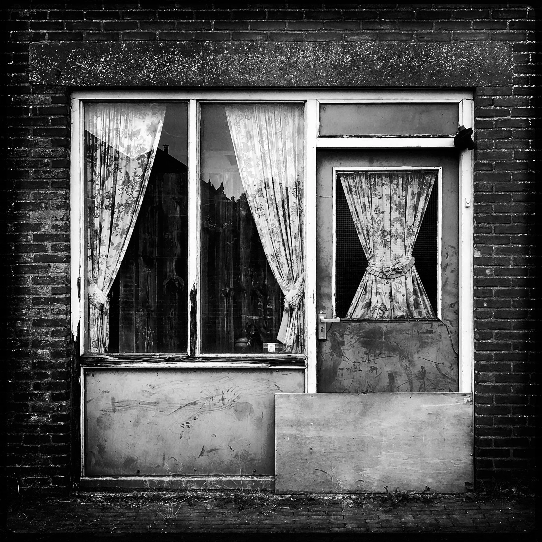 Black and white photo of a weathered building with a large window and a door, both covered with floral curtains. The building appears abandoned.