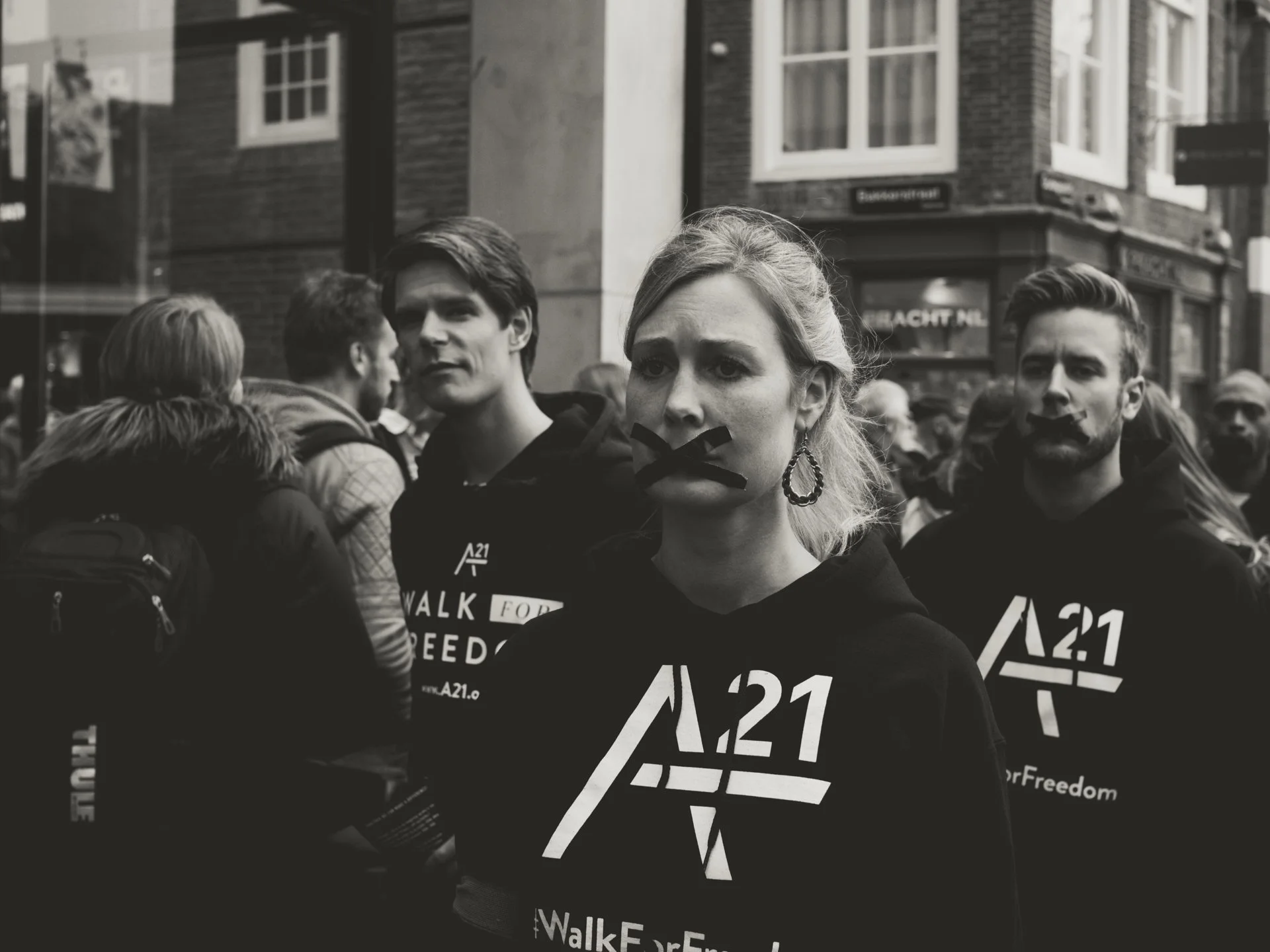 People wearing black hoodies with 'A.21' and 'Walk for Freedom' text, protesting on a city street, with buildings and other protesters in the background.