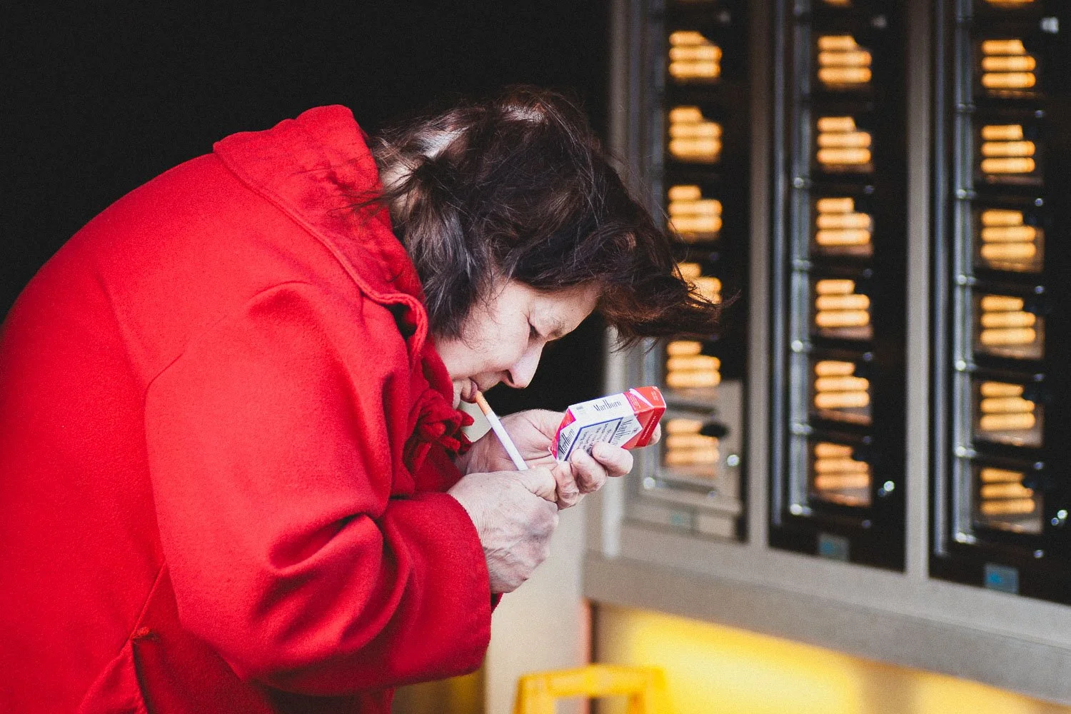 A woman in a red coat is smoking a cigarette while holding a small carton of milk or juice in her other hand. She is leaned forward near a vending machine with snacks.