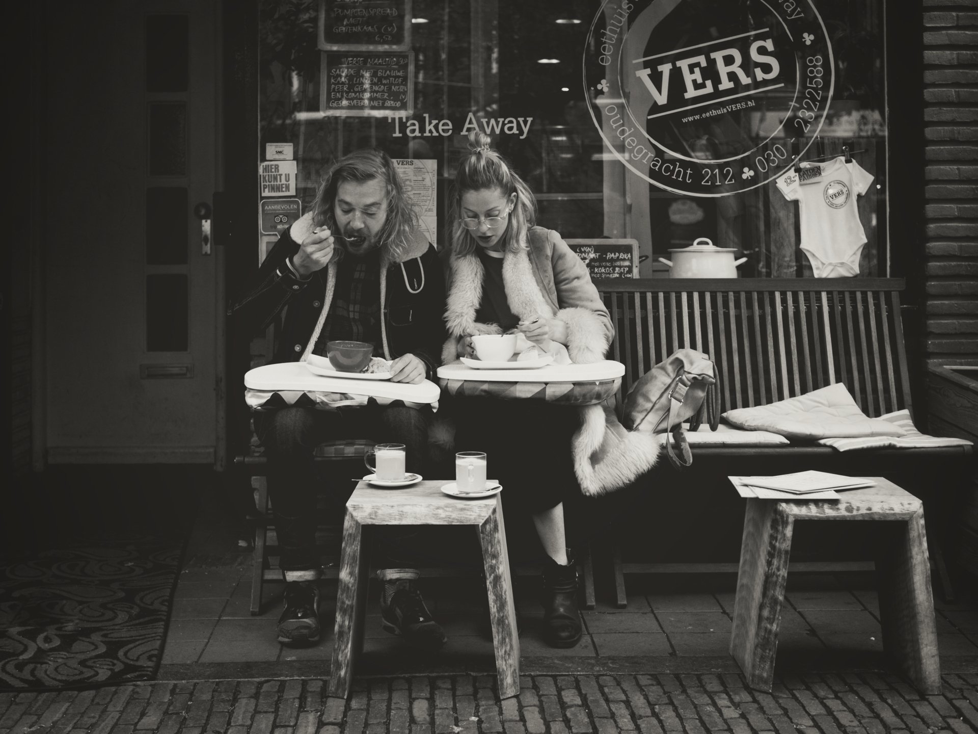 Two women sitting at an outdoor cafe table, eating and drinking. One woman has a fork near her mouth, and the other uses a spoon, with cups and plates on the table. A bench with a coat, a small table with cups, and a cafe window with signs and a smal