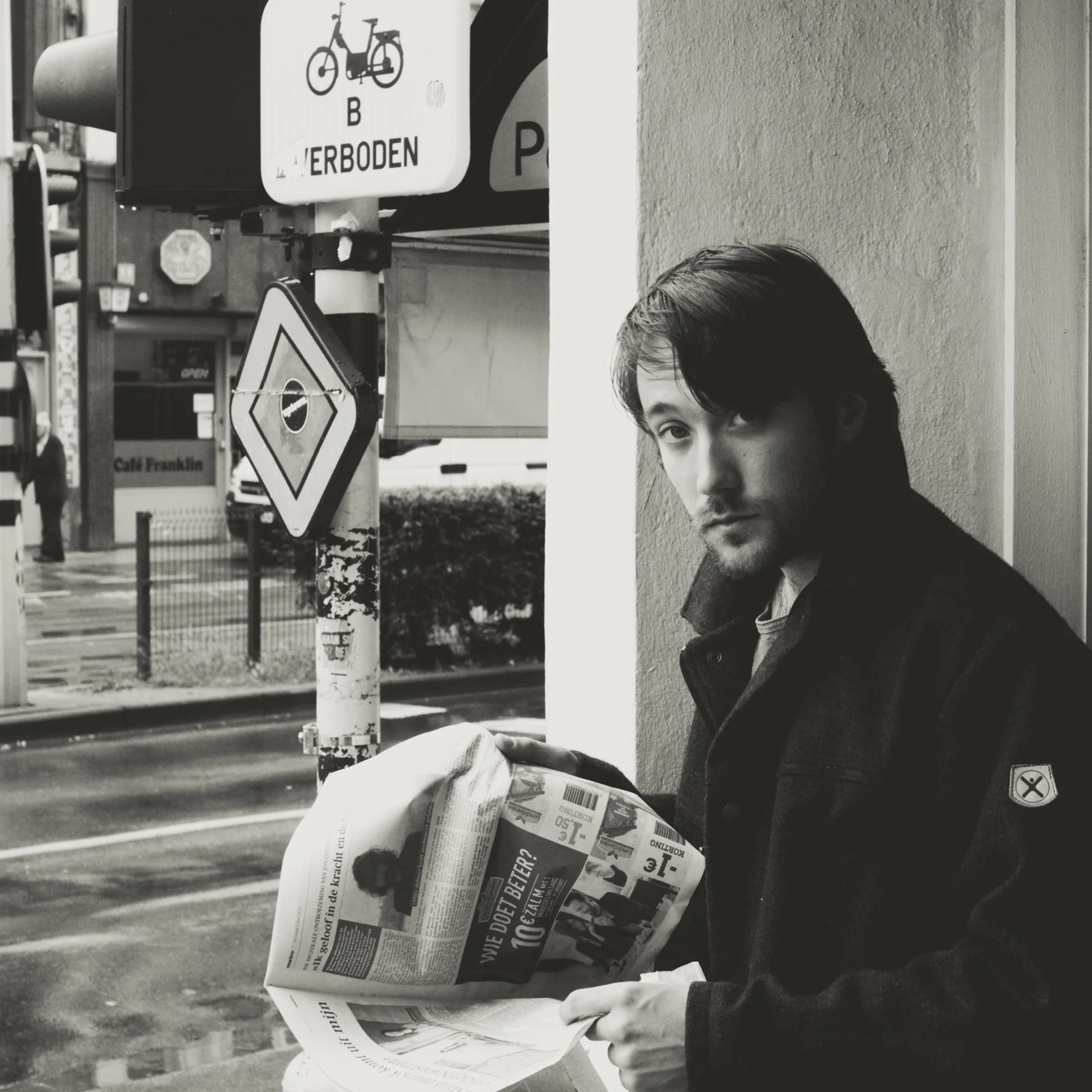 A young man with dark hair and a beard holding a newspaper outside on a city street. There are traffic signs and a wall behind him, with a person walking in the distance.