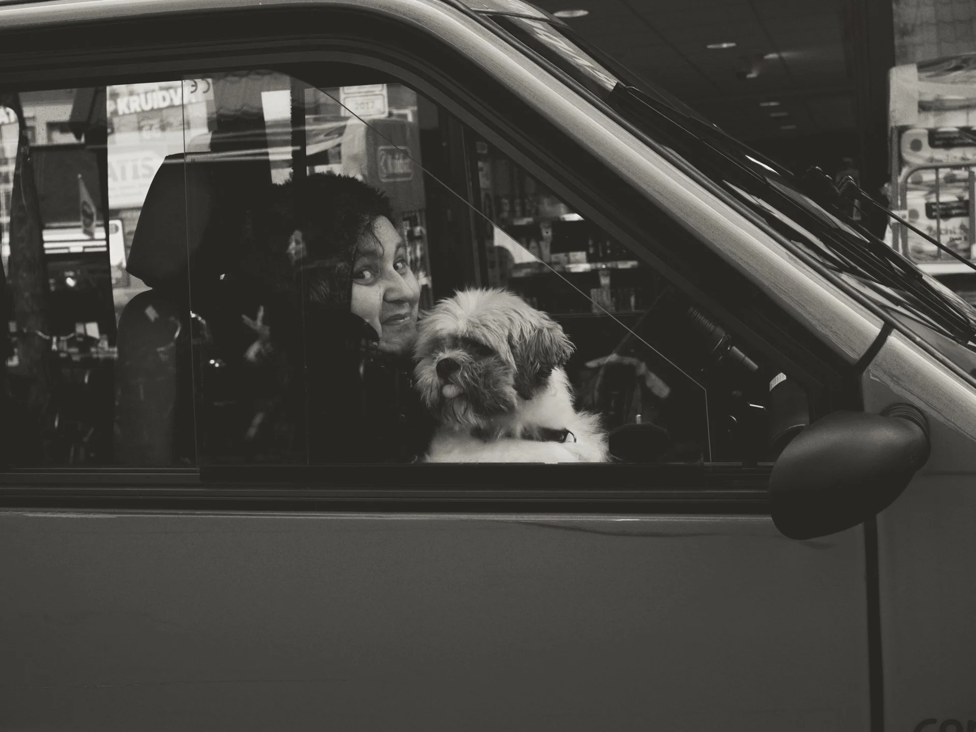 Woman with curly hair smiling with a small dog inside a vehicle, viewed through the window, with a busy street and stores in the background, in black and white.
