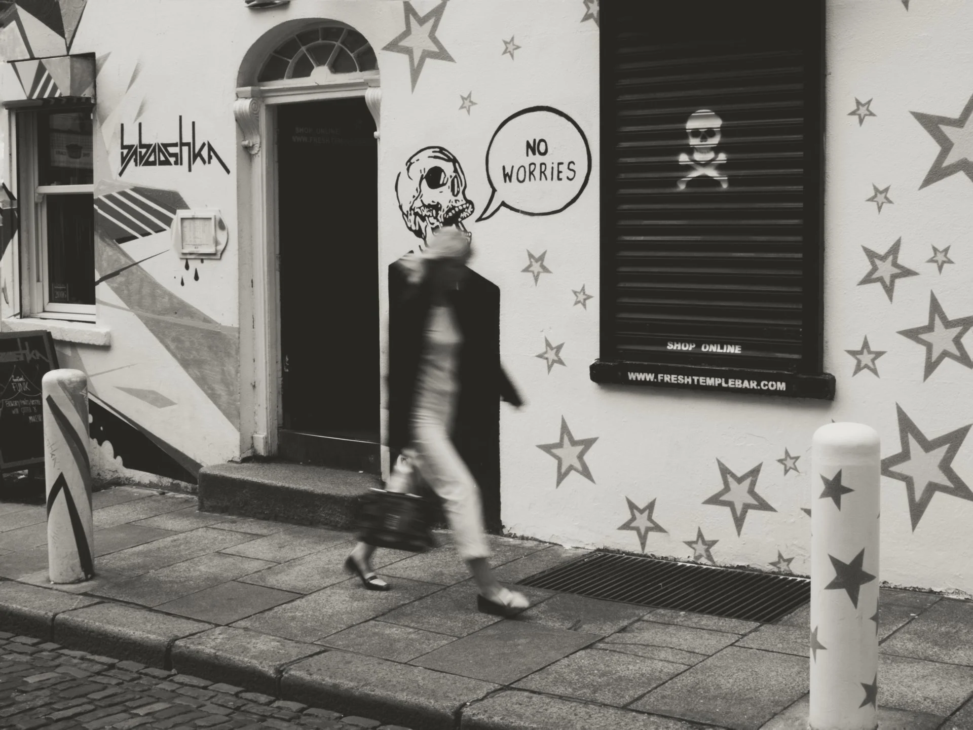 Black and white photo of a man walking past a building with graffiti art. The graffiti includes a skull with a speech bubble saying 'NO WORRIES' and a skull and crossbones on a shutter. The wall is decorated with star and geometric designs. There are