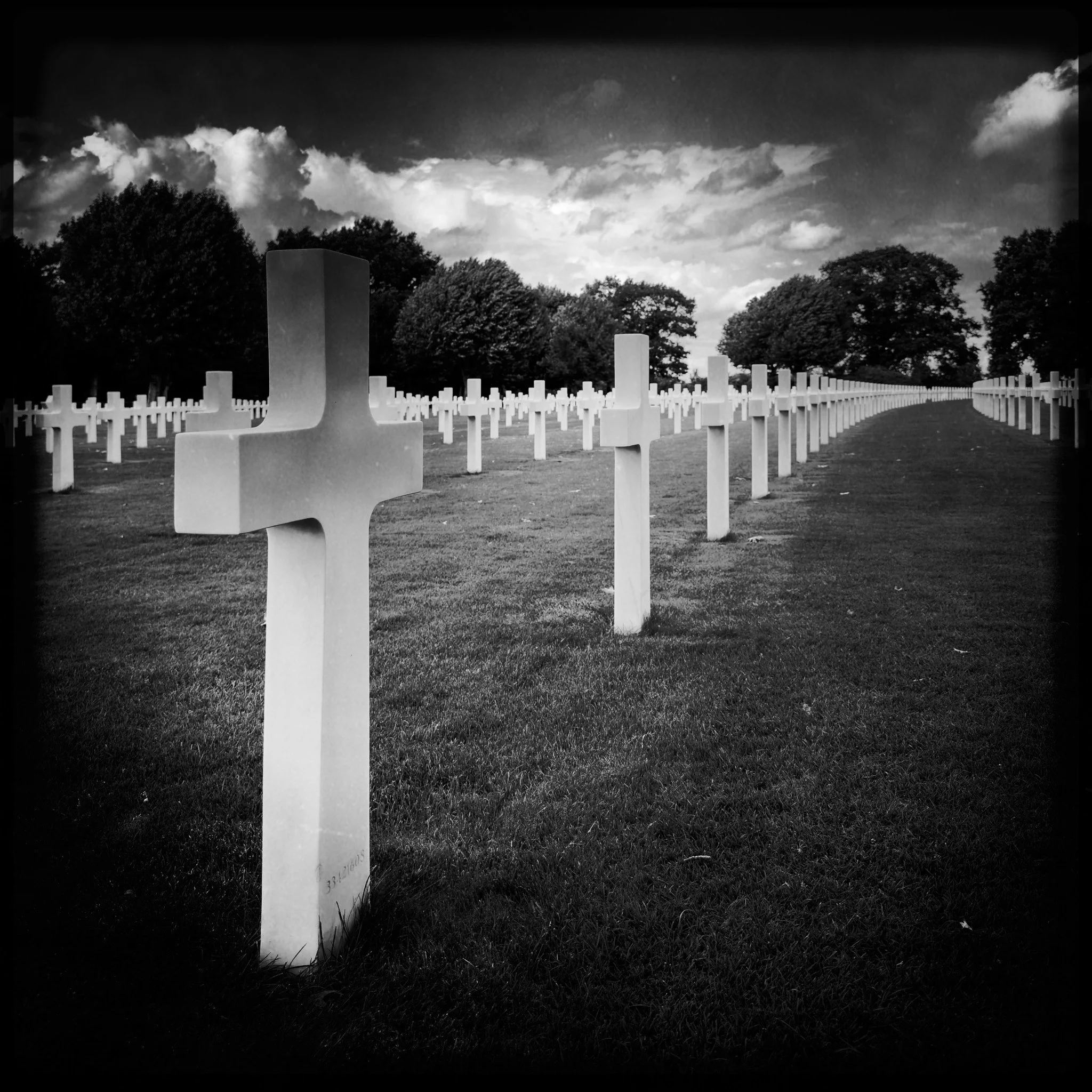 Black and white photo of a military cemetery with rows of white crosses on a grassy field, trees in the background, and cloudy sky overhead.