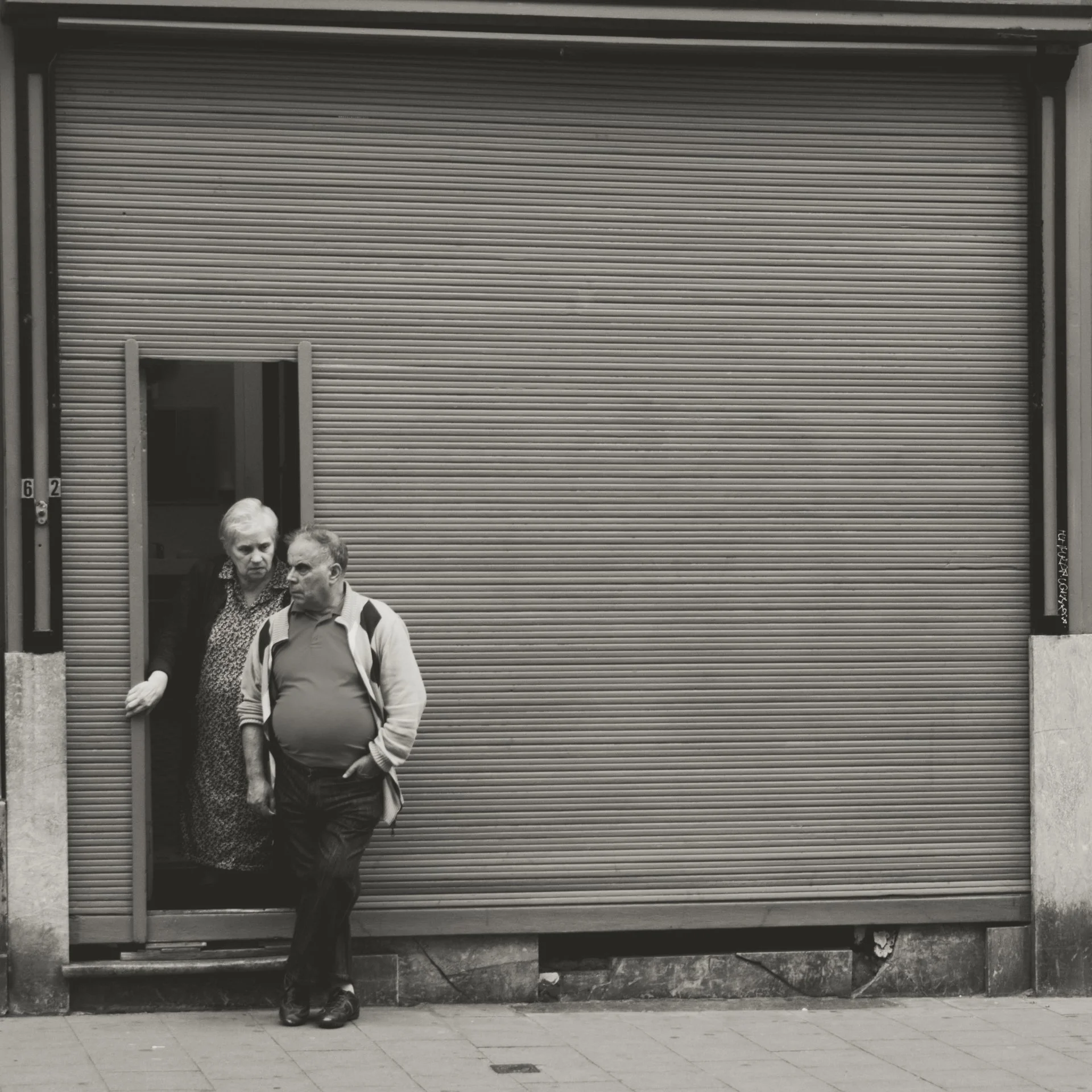 Two elderly women and one elderly man walking on a sidewalk in front of a closed metal roll-up door. The women are engaged in conversation, while the man appears to be walking alone.