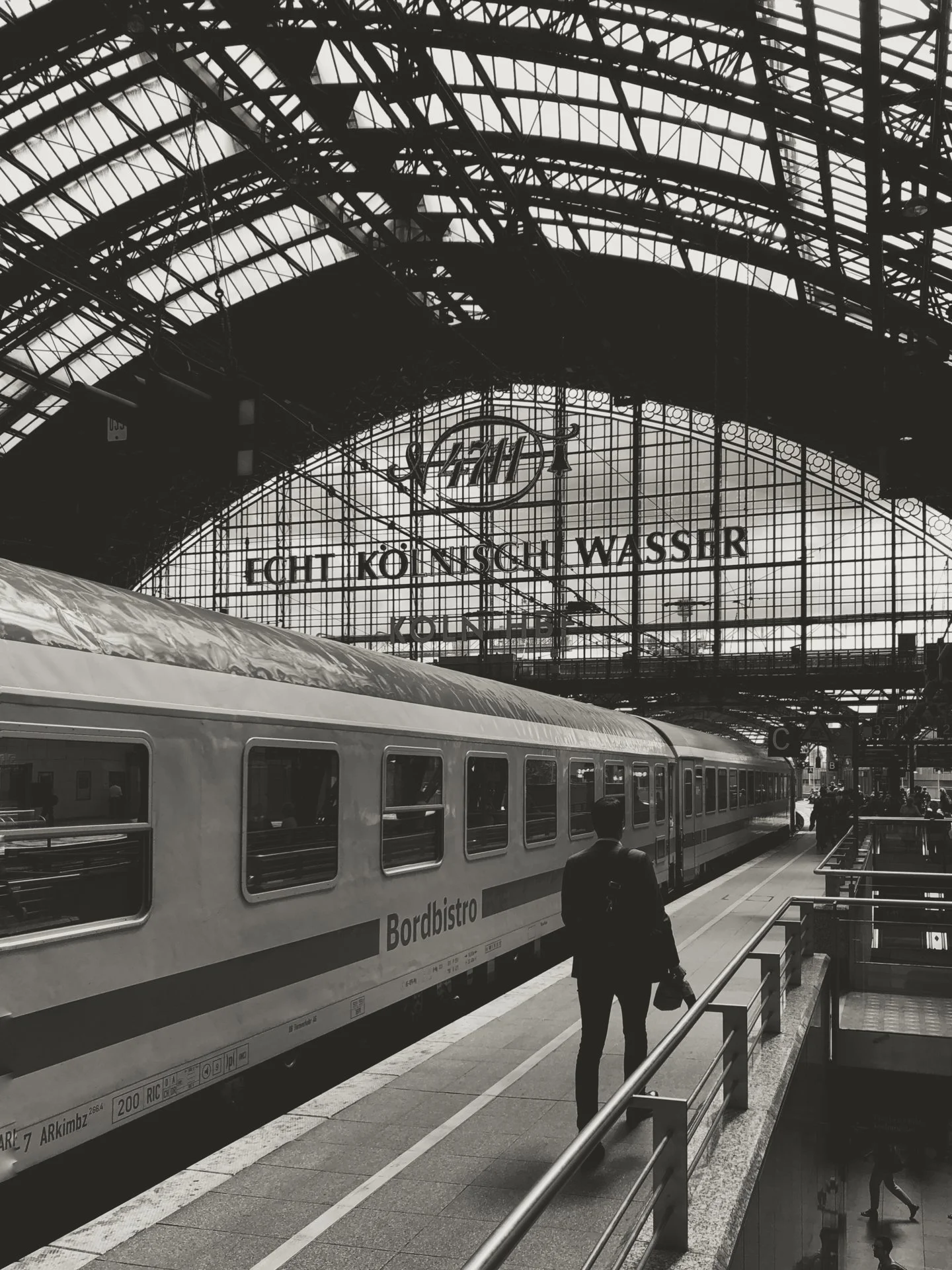 Black and white photo of a train station with a train on the platform and a large glass roof overhead. A man with a backpack is walking along the platform. The station has a sign that reads 'Licht Kölnisch Wasser' and the logo 'FCH' is visible.