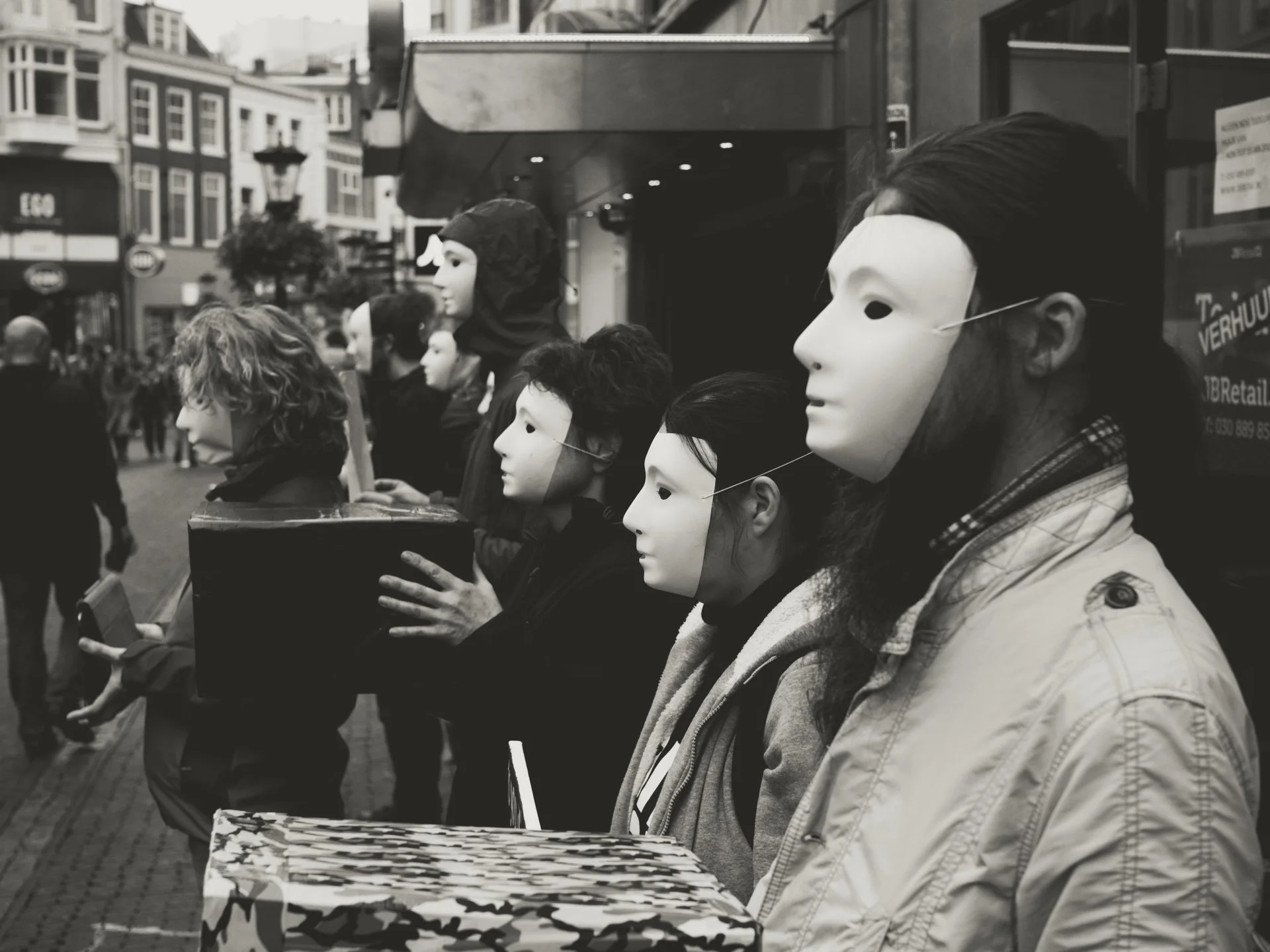 People wearing masks standing in a line on a street, possibly participating in a protest or event, with buildings and pedestrians in the background.