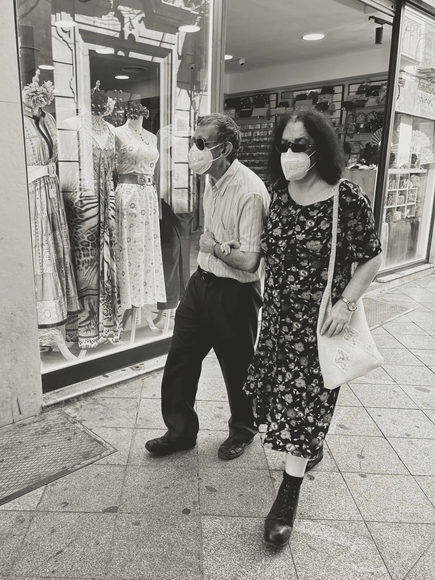 Two women walking past a store window displaying mannequins in dresses. Both women wear face masks and sunglasses, one woman in a floral dress and the other in a striped shirt and dark pants. They are carrying bags and walking on a sidewalk.