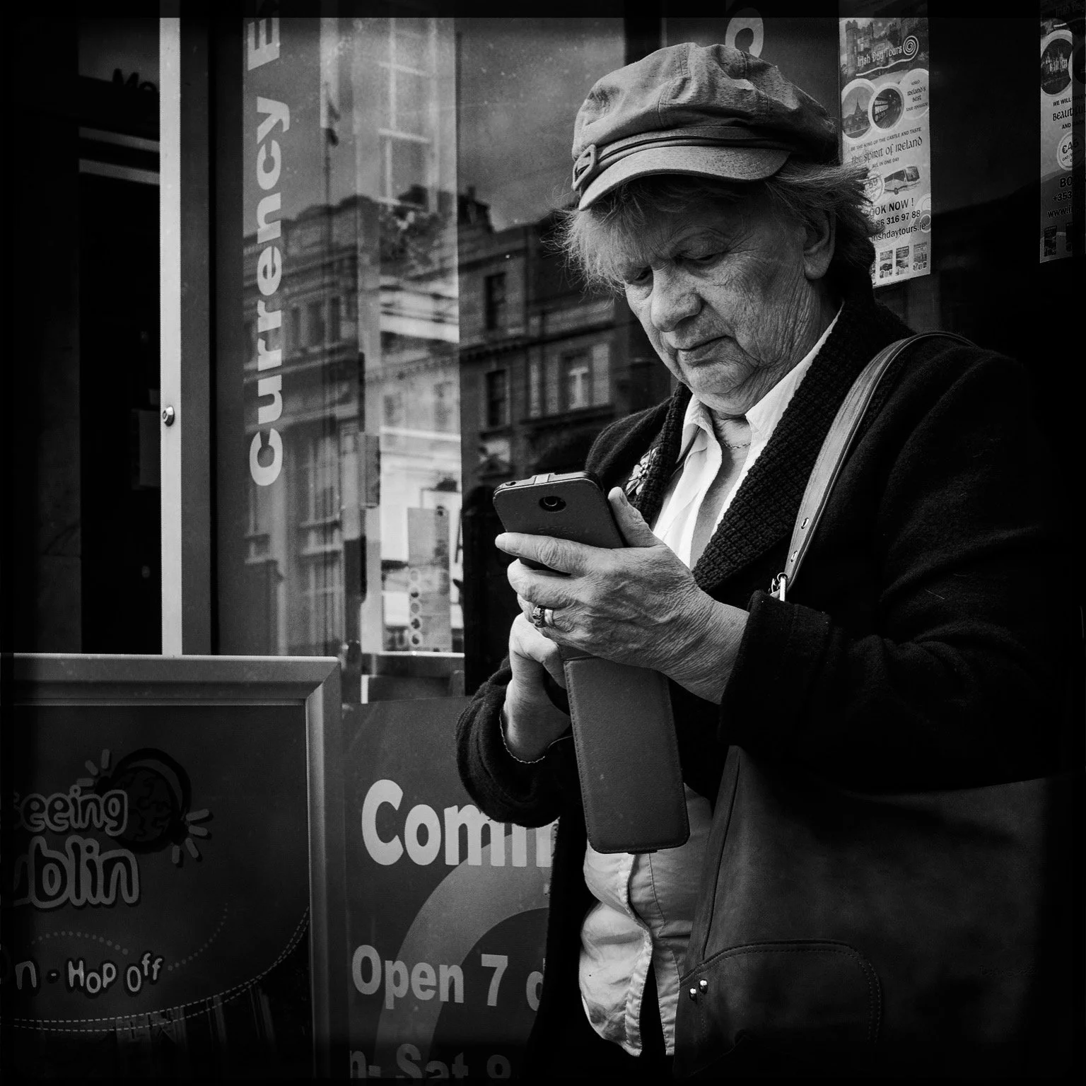 An elderly woman wearing a hat and a dark coat is looking at her phone while standing outside a store. Reflection of buildings is visible on the store window.
