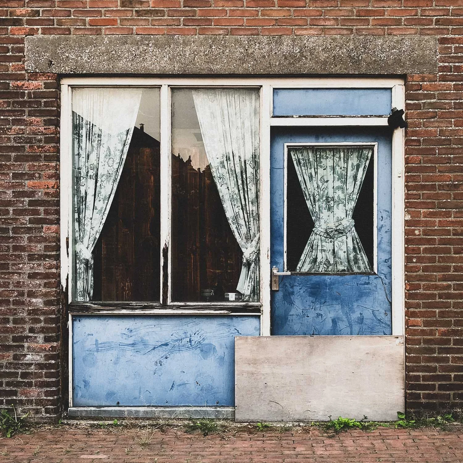 An old brick building with a blue door and windows, floral curtains, and a boarded-up lower section.