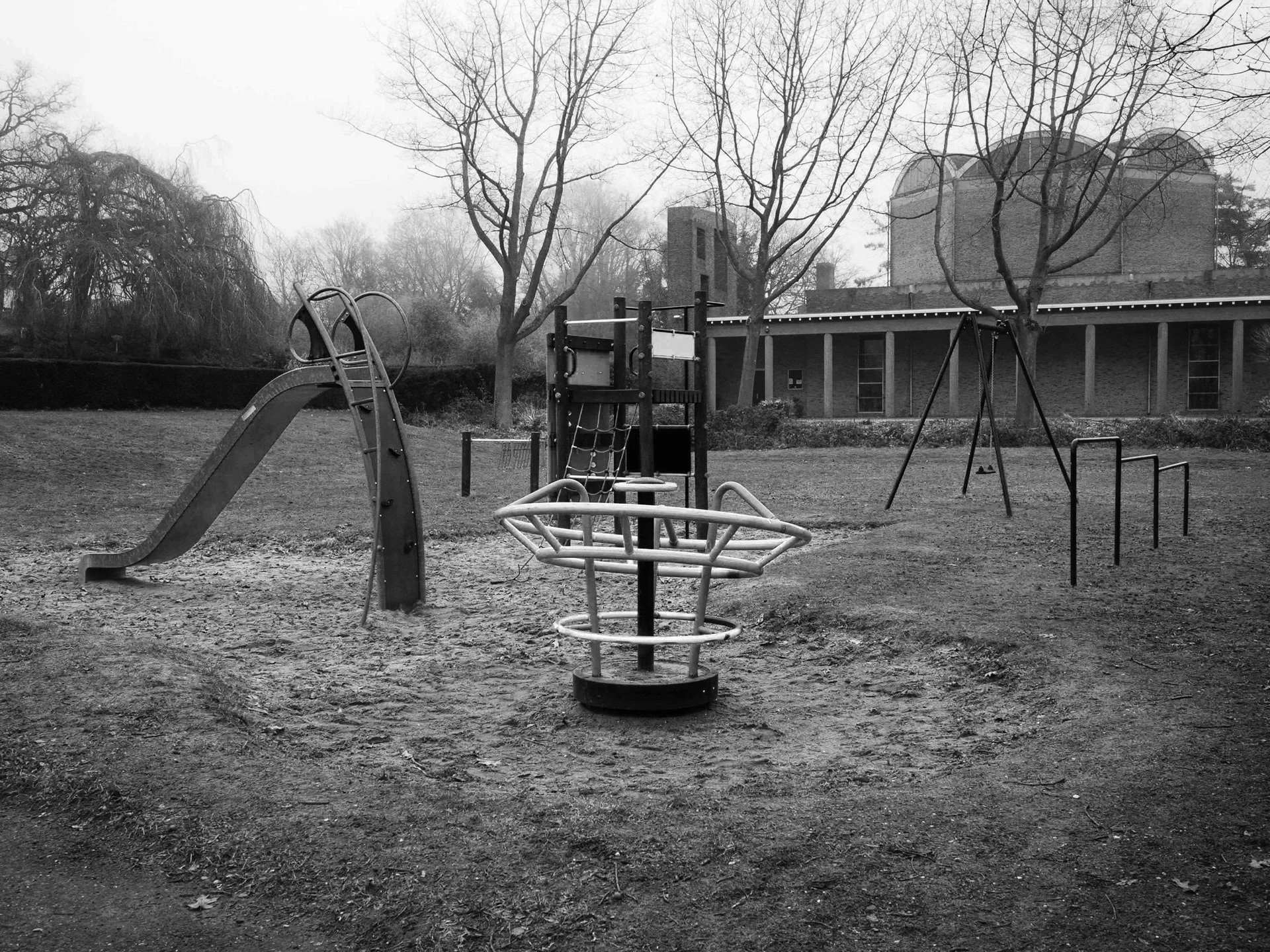 Empty playground with slide, swings, and merry-go-round, surrounded by leafless trees and a building in the background, in black and white.
