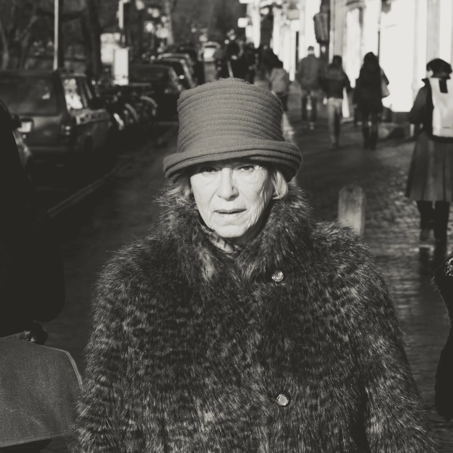 Close-up of an elderly woman wearing a large knit hat and a furry coat, walking on a busy city sidewalk with cars parked on the side and pedestrians in the background.