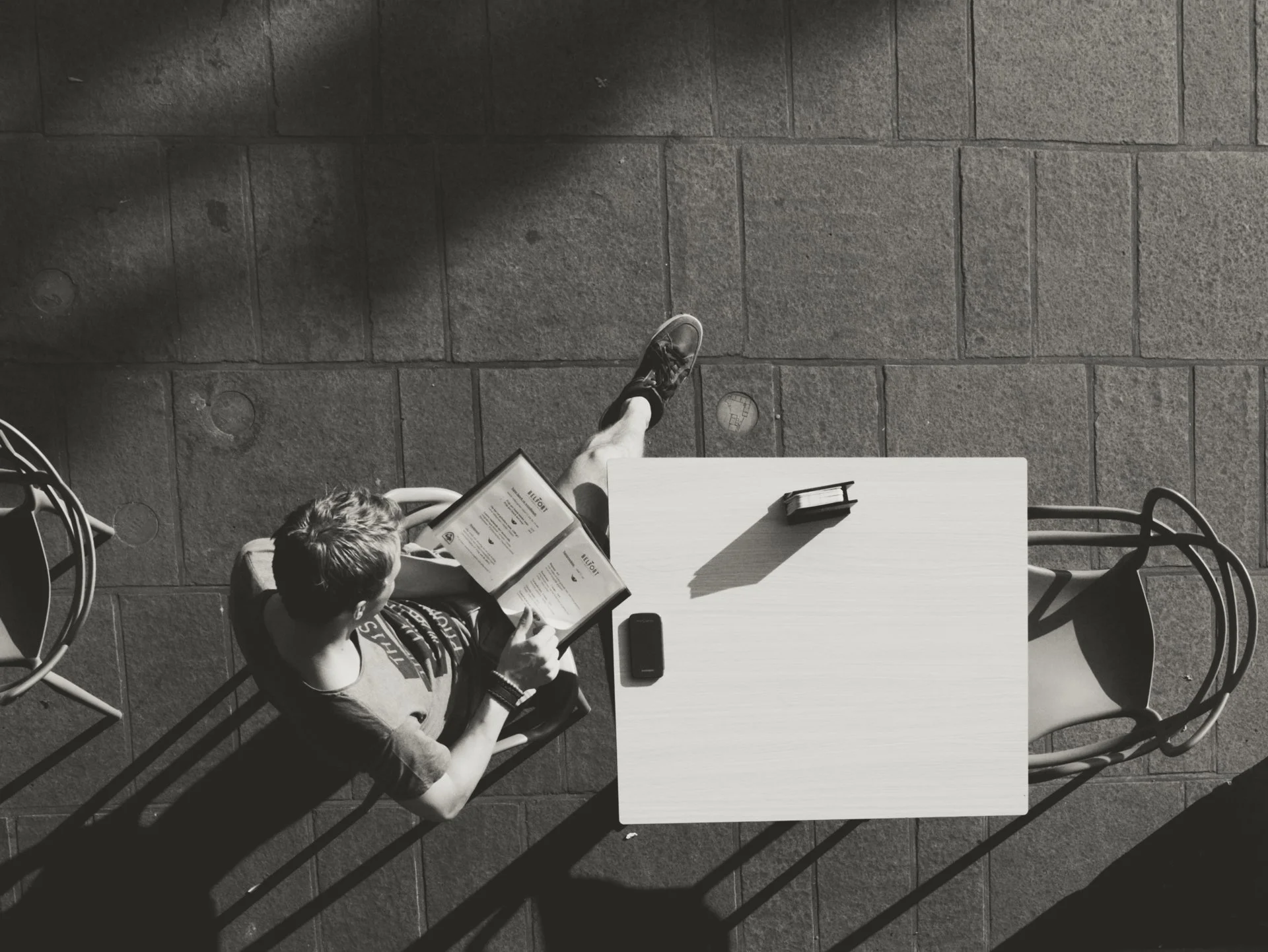 Black and white photo of a person sitting at an outdoor table, reading a menu, with two mobile phones on the table. Shadows of a railing are cast on the ground.