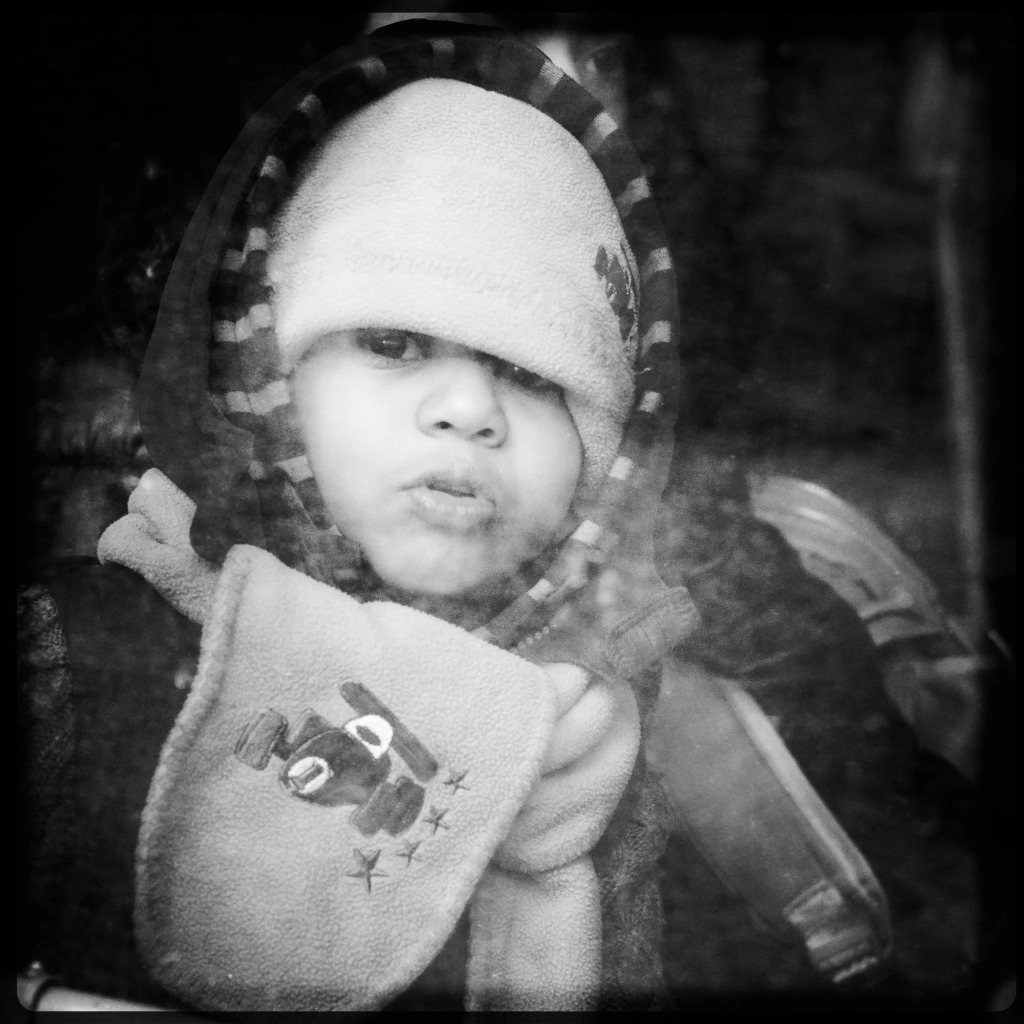 A young child looking through a glass window, wearing a hoodie and a hat, with a scarf covering part of his face and holding a piece of cloth with the word "HOPE" on it.