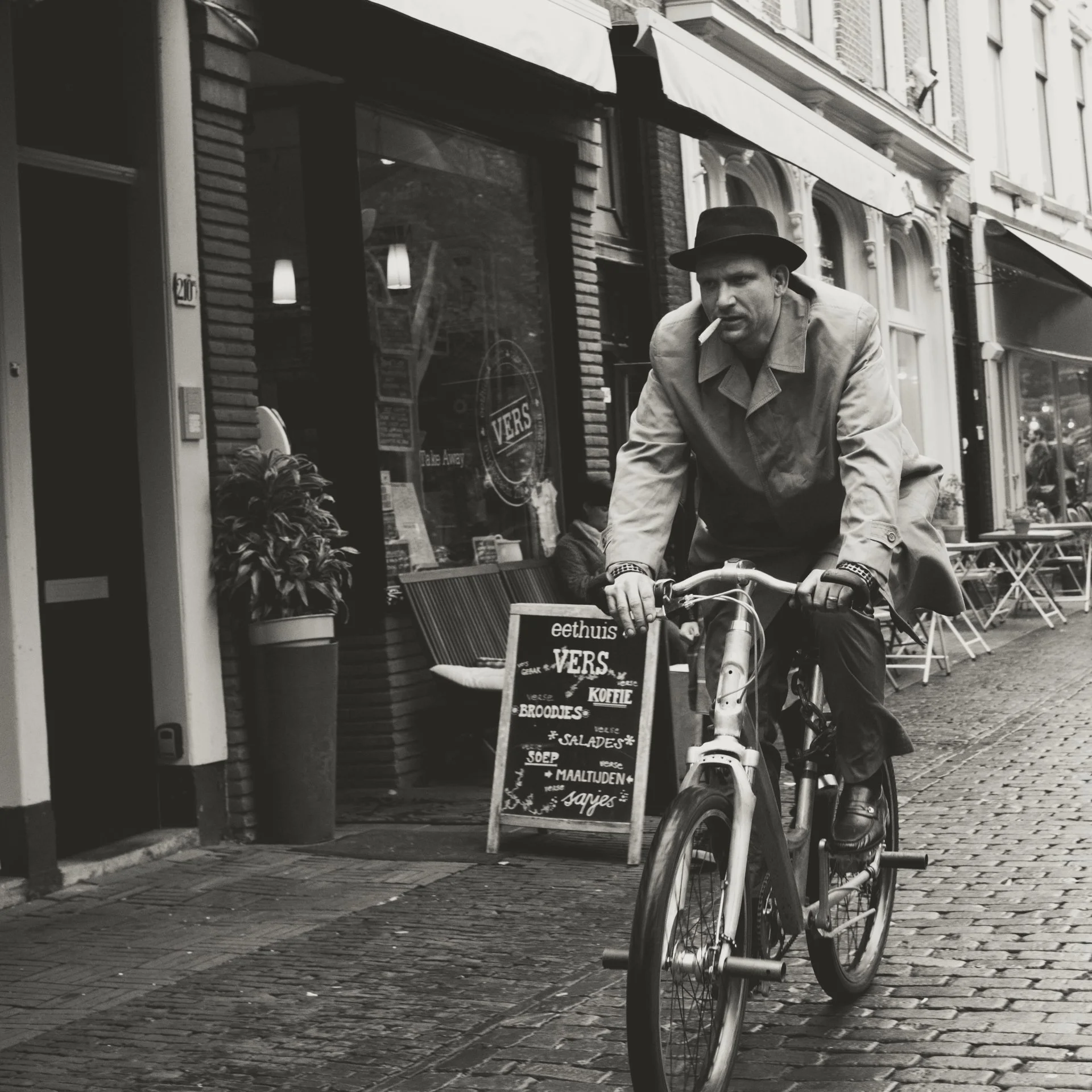 A man wearing a fedora hat and a trench coat rides a bicycle on a cobblestone street in an urban area. There is a storefront with a sidewalk sign listing menu items such as coffee, croissants, bread, salads, and soup. The background includes tall bui