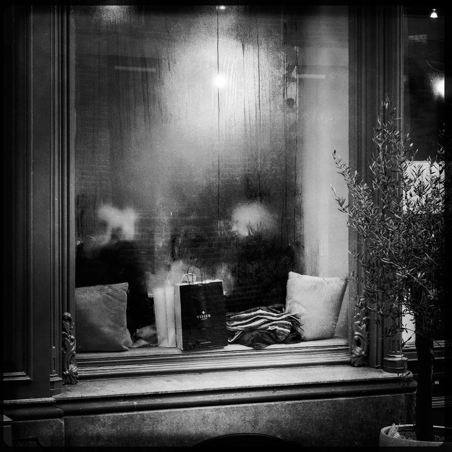 A black and white photo of a window display with two pillows, a shopping bag, some folded fabric, and a potted plant outside.