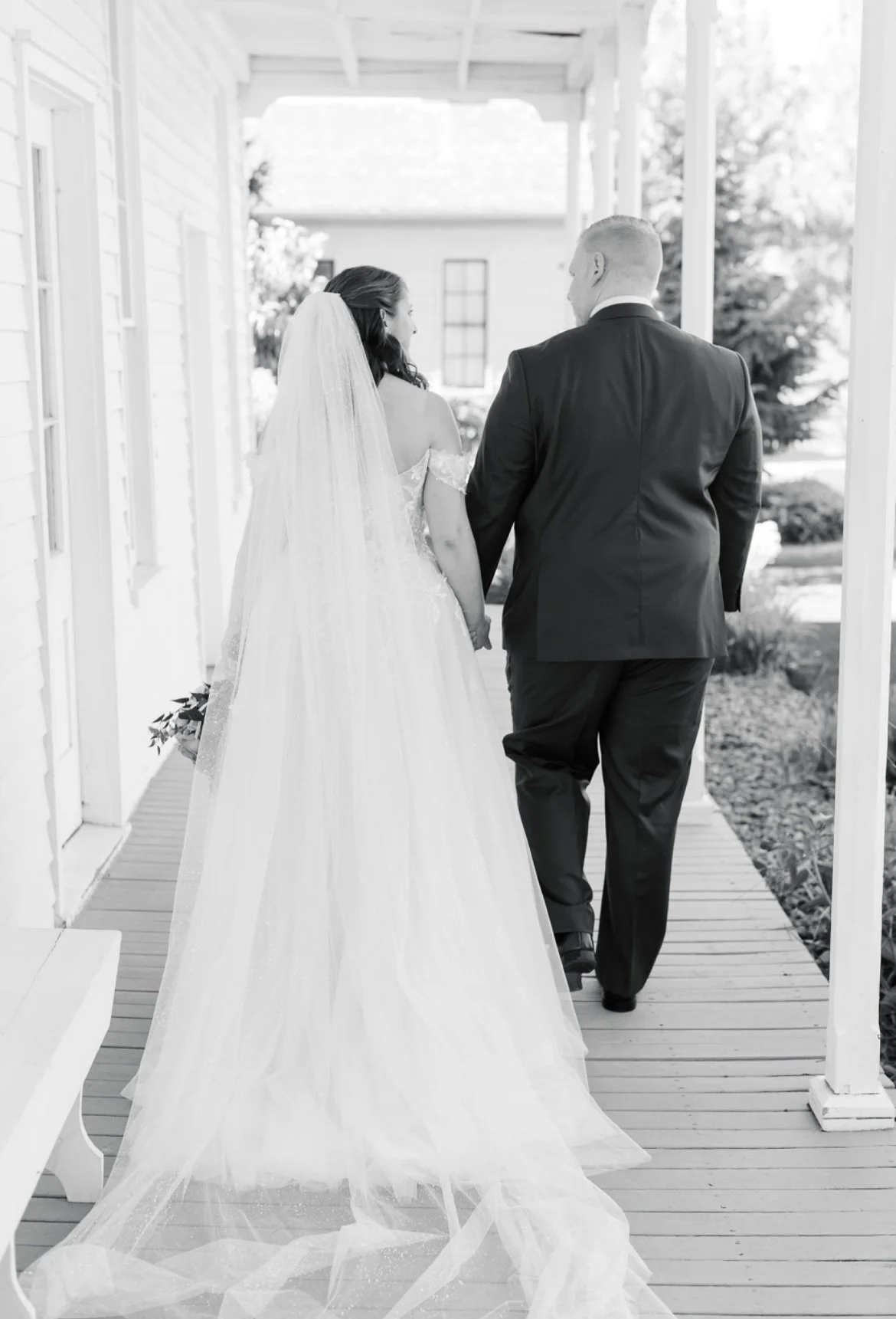 Bride and groom walking hand in hand on a porch, bride wearing a long veil and gown, groom in a suit, black and white photo.