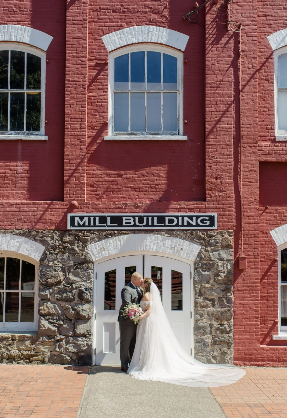 A couple in wedding attire kissing in front of a red brick building with arched windows and a sign that reads 'Mill Building.'