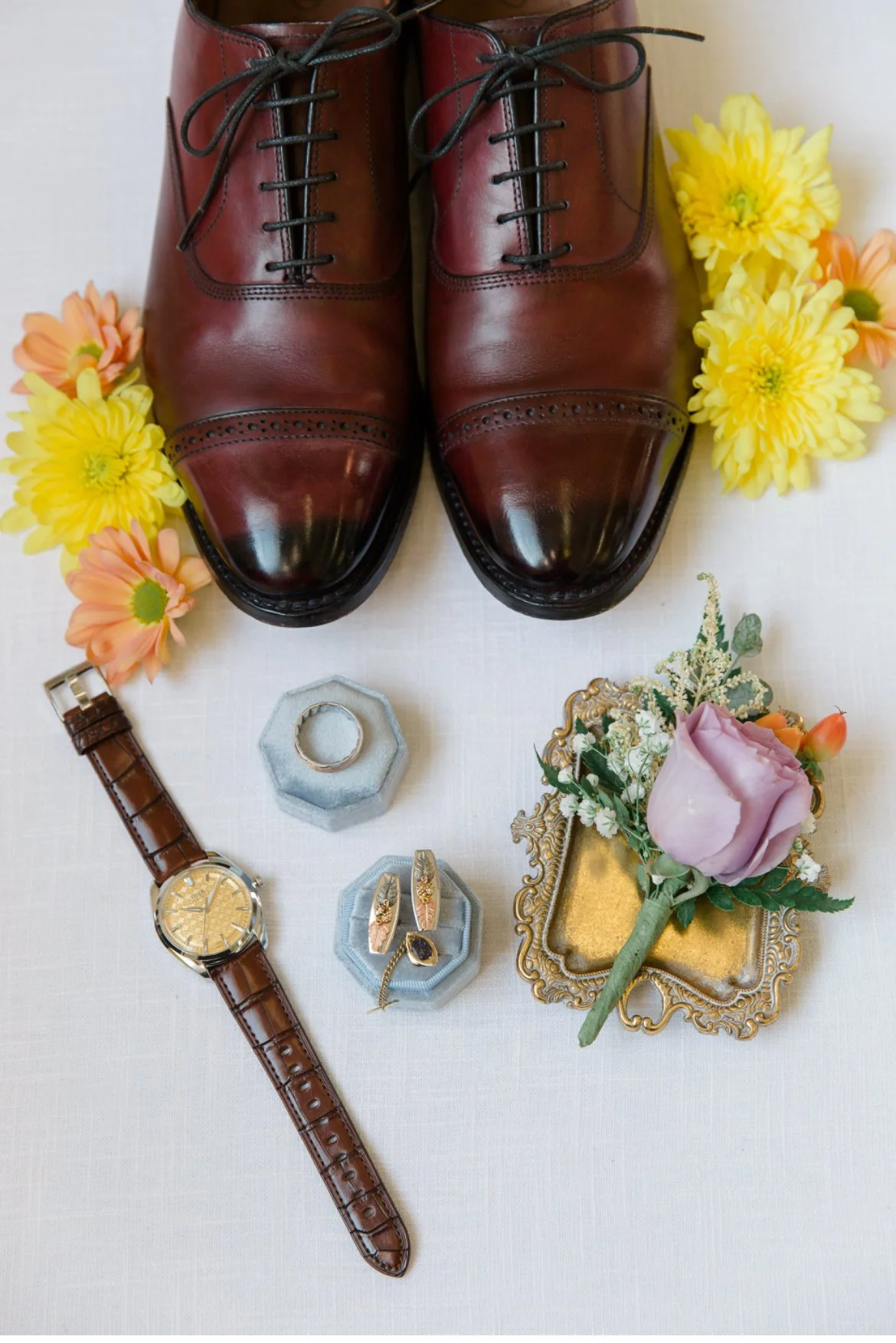 Brown dress shoes with lace, surrounded by yellow and orange flowers, a wristwatch with a brown leather band, a wedding ring in a blue box, cufflinks, a vintage tray, and a boutonniere with a purple rose.