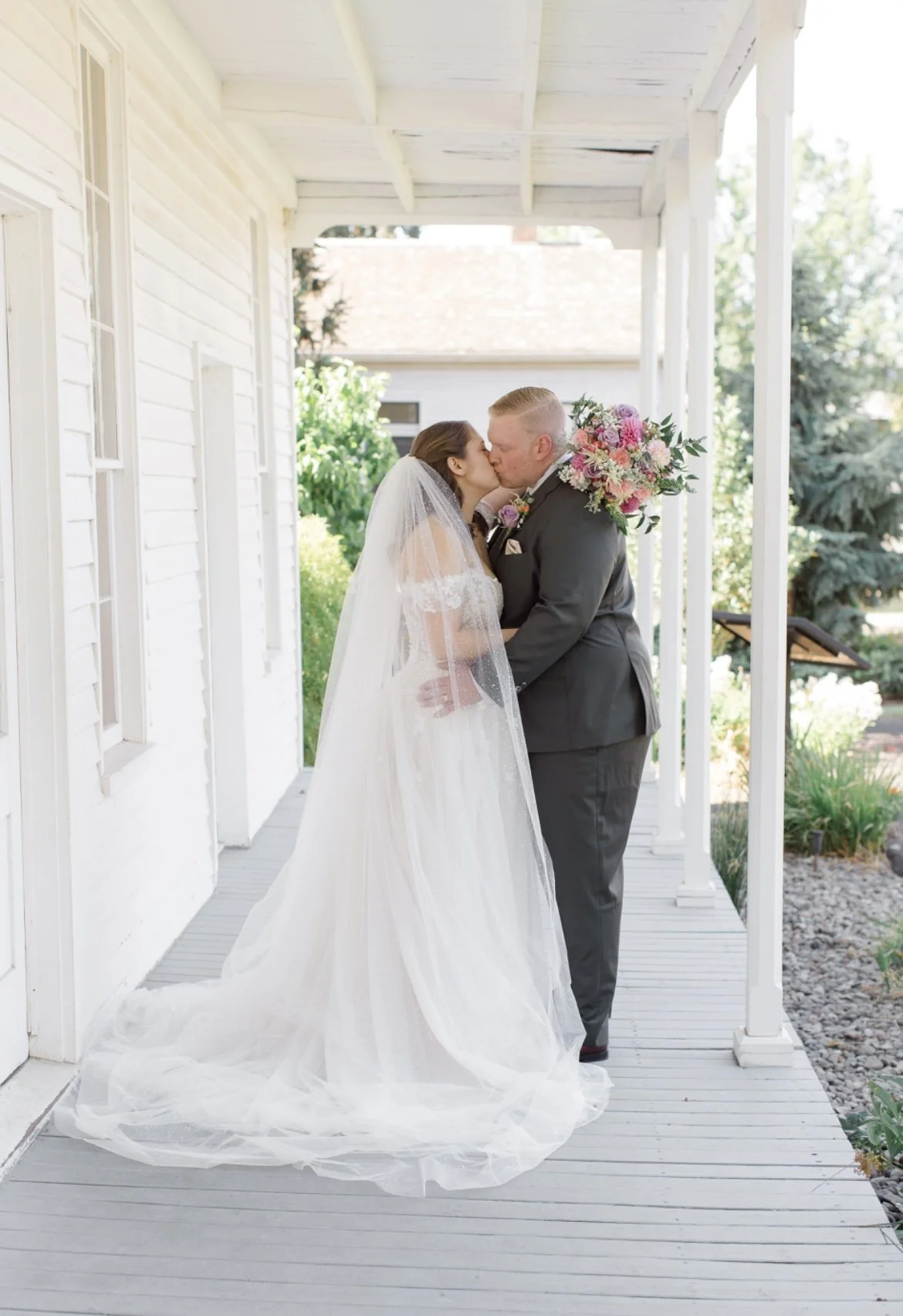 Bride and groom kissing on a porch, bride in a white gown and veil, groom in a black suit holding a colorful bouquet.