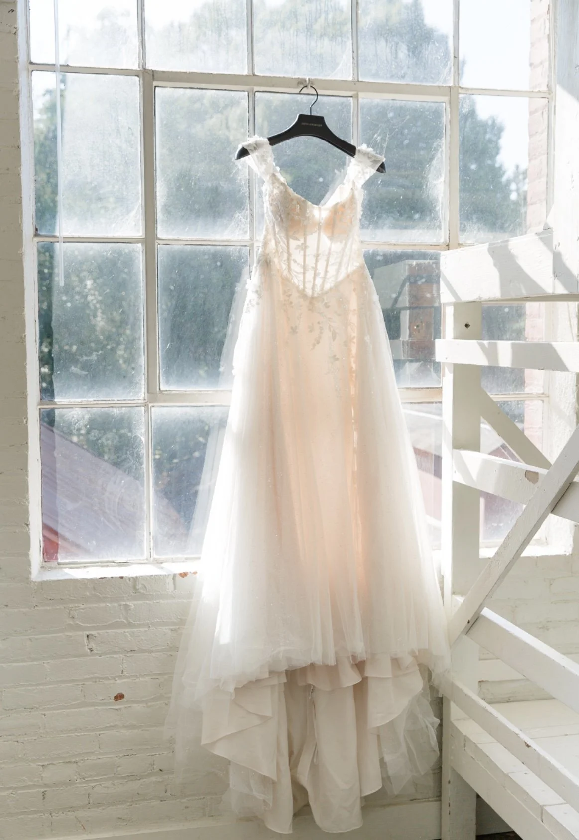 White wedding dress hanging on a black hanger in front of a large window, with sunlight streaming in.