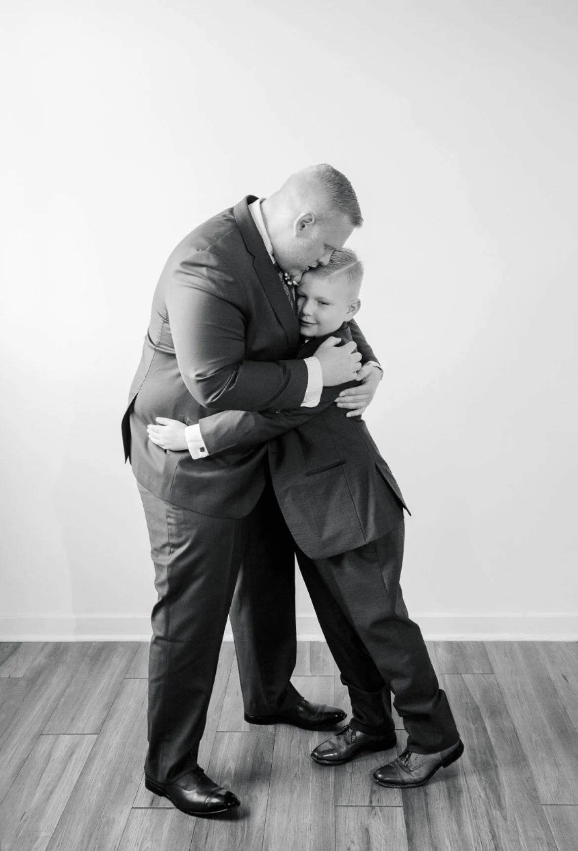 Black and white photo of two people in suits hugging, likely a father and son, on a wooden floor.