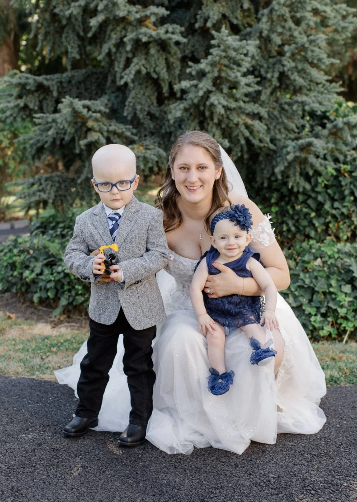 A smiling woman in a white dress crouches next to a young boy in a gray blazer and glasses holding a toy, and a baby in a blue dress with a headband.