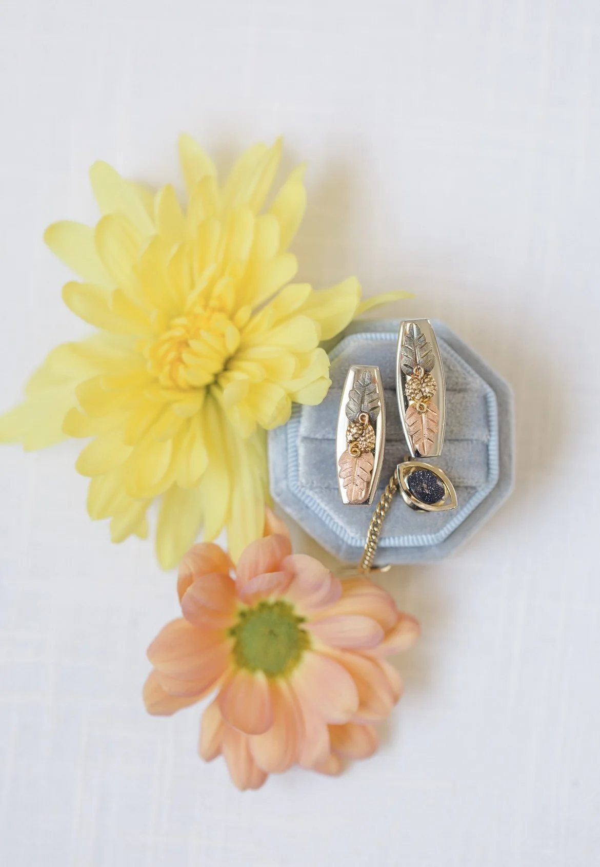 Two floral-design earrings and a ring with a dark stone on a light blue jewelry box, surrounded by yellow and peach flowers.
