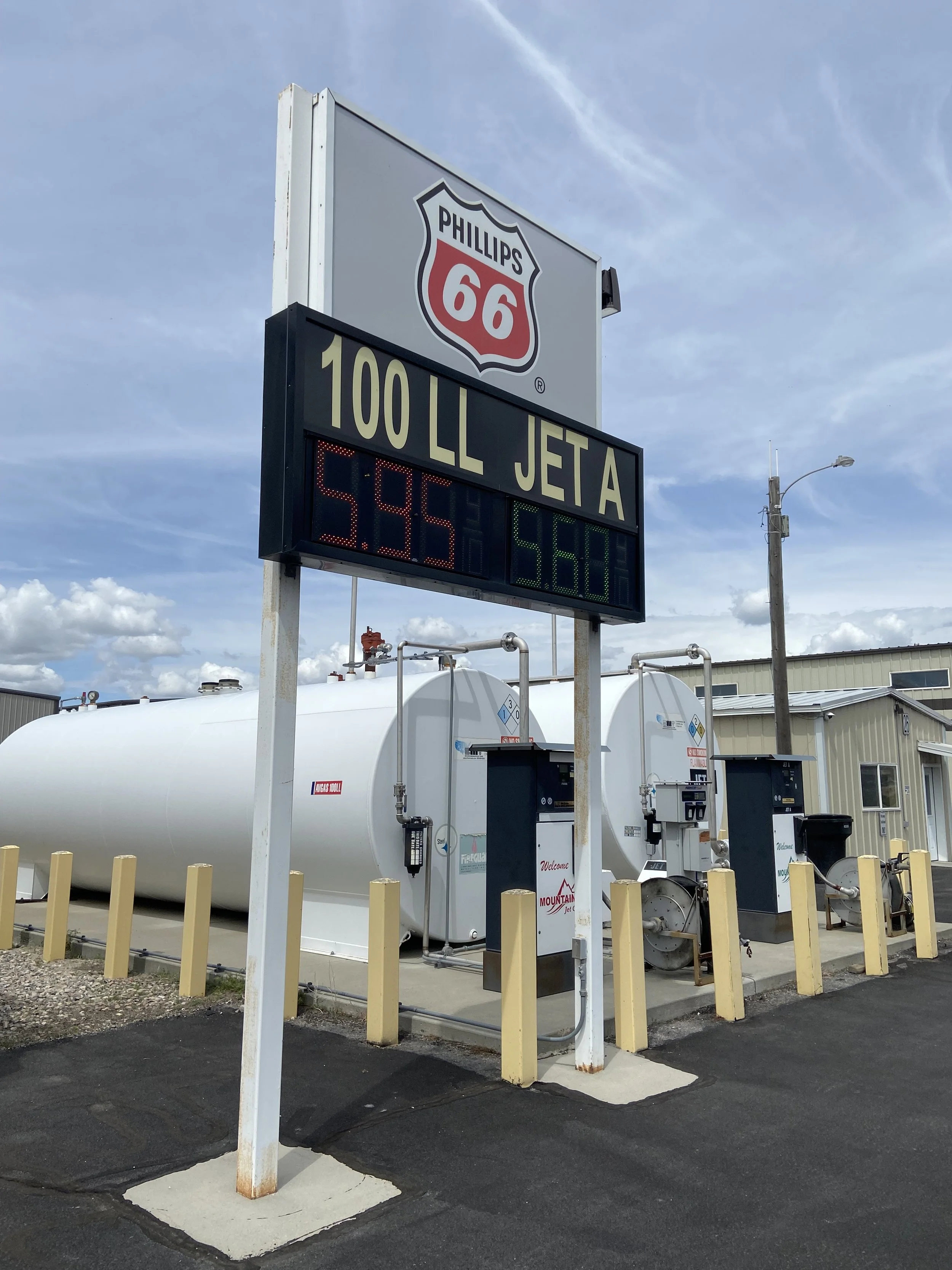 Gas station signage displaying fuel prices, with large propane tanks and industrial buildings in the background under a partly cloudy sky.